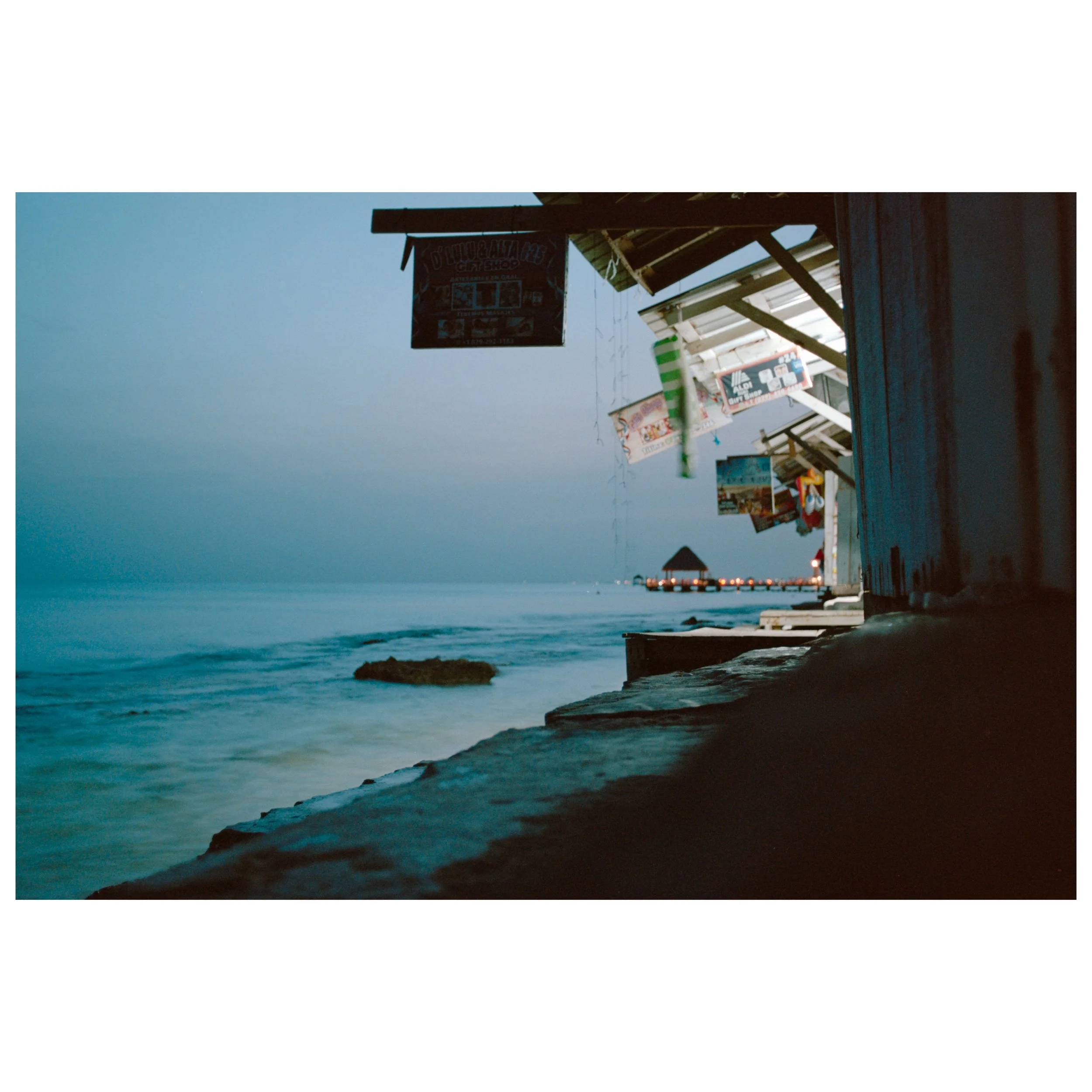 Beachside shops and huts at dusk, with calm ocean waves and distant pier with lights.