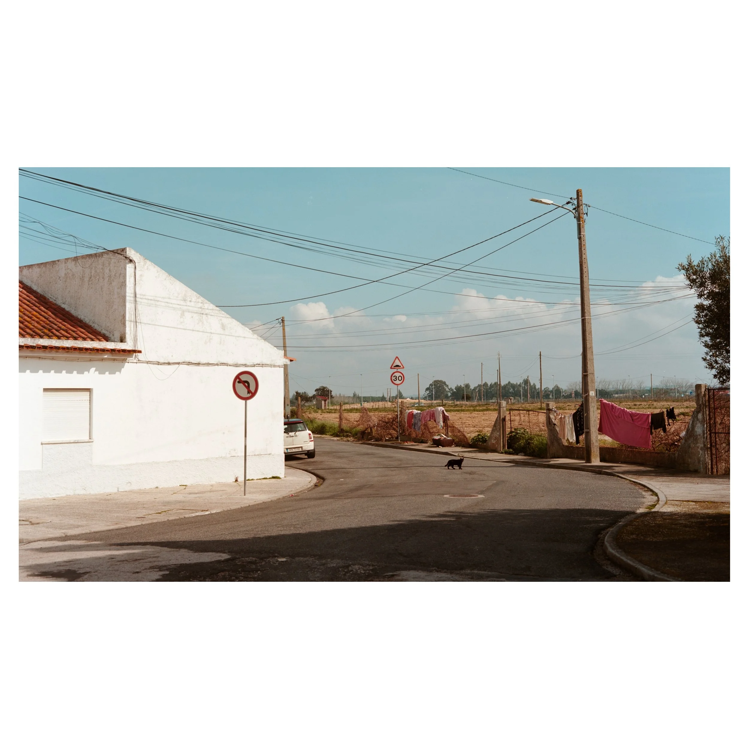 A quiet street corner with a white building on the left, a no left turn sign, and a car parked along the curb. Power lines crisscross the sky, and laundry hangs on a fence on the right side of the street. The background features open fields with a fe
