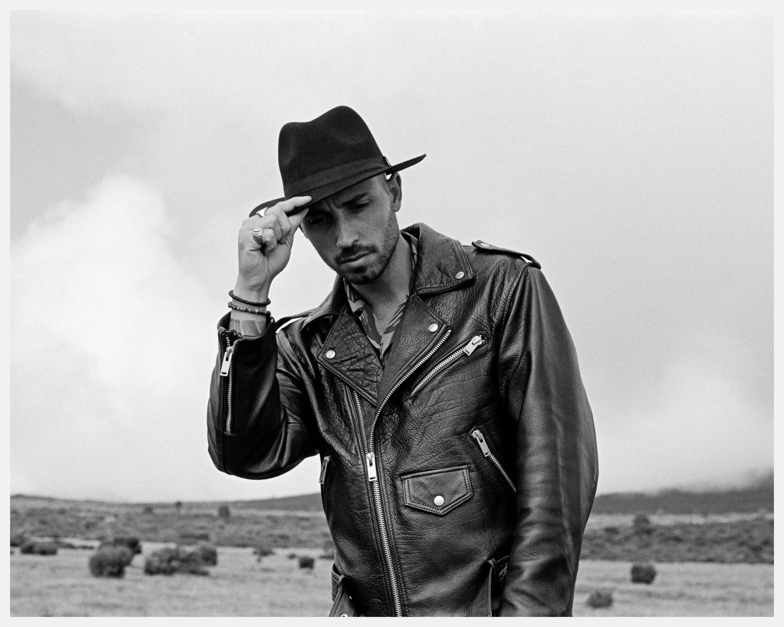 A man wearing a leather jacket and a fedora hat, standing outdoors in a field, looking at the camera with a serious expression.