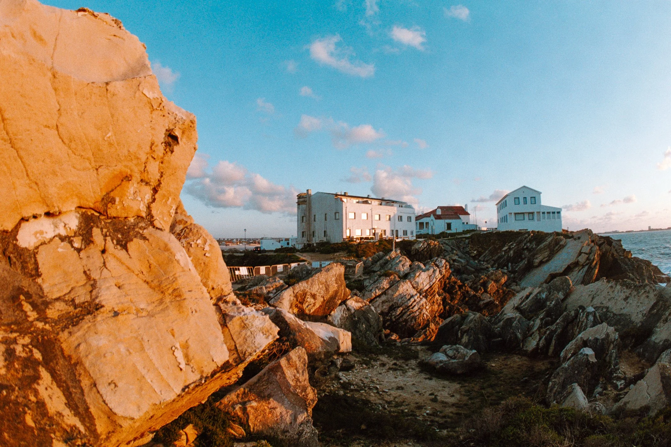 Rocky coastline with large beige and dark rocks, white houses with red roofs, and a blue sky with clouds at sunset.