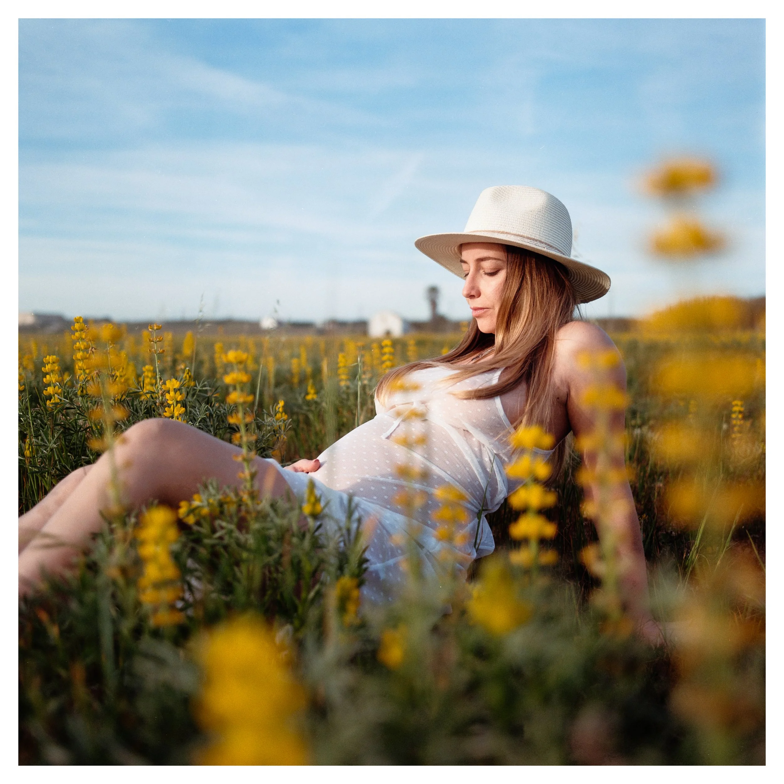 A woman in a white dress and wide-brimmed hat relaxing in a field of yellow flowers under a blue sky. photo by Bruno Novais