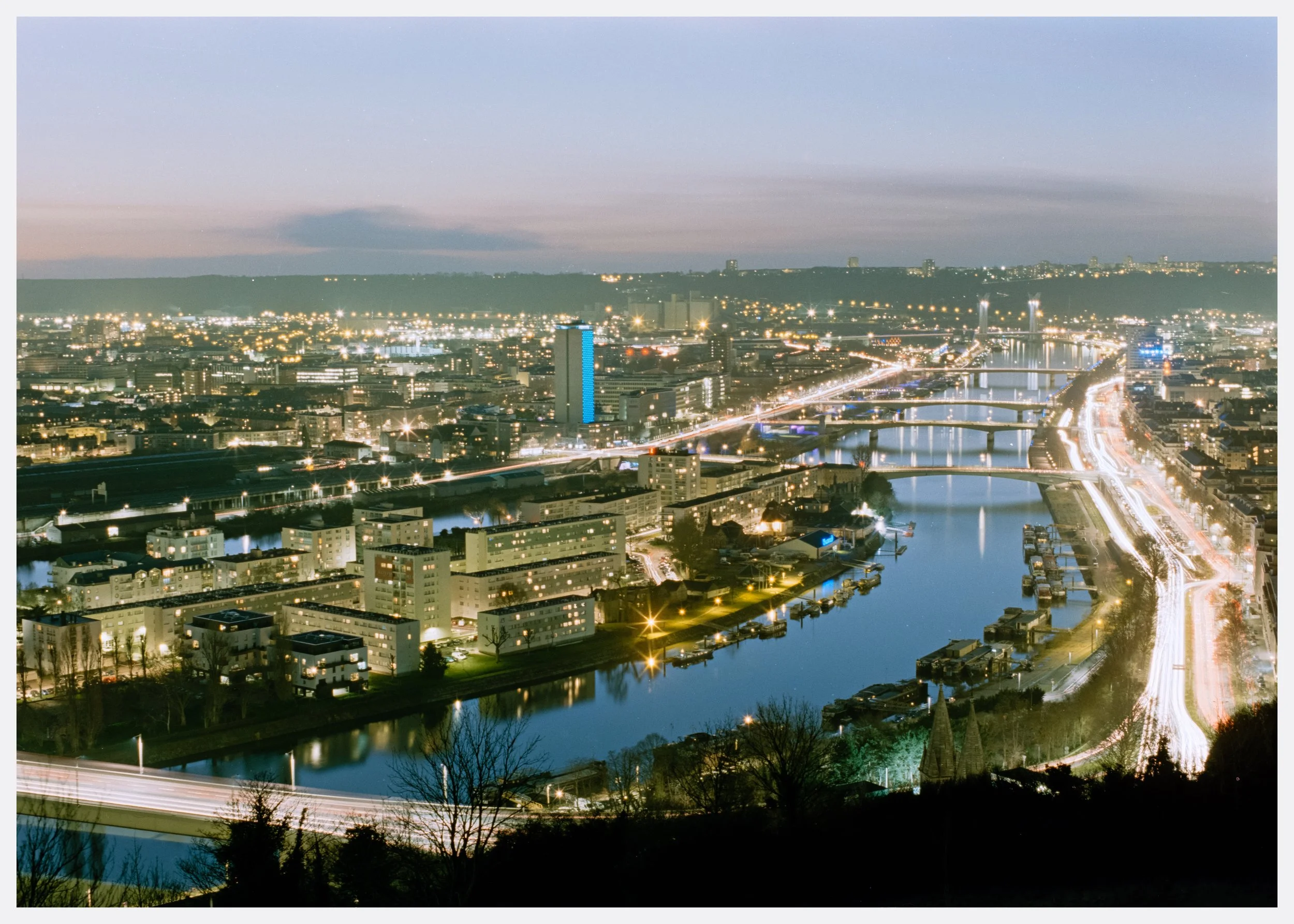 Nighttime cityscape with illuminated buildings, bridges, and a river showing reflections, with light trails from moving vehicles.