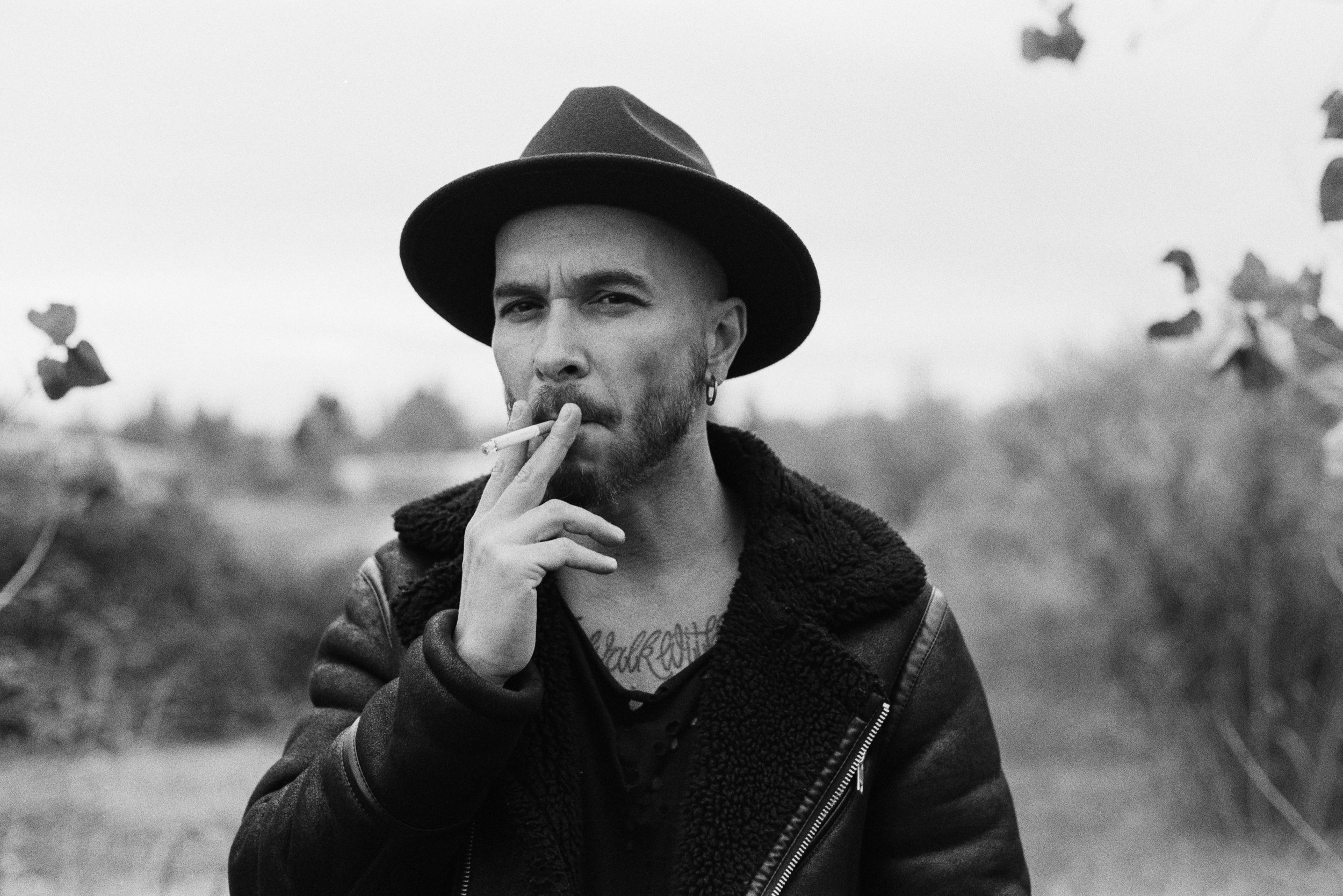Black and white photo of a man wearing a fedora hat, leather jacket with shearling collar, and earrings, smoking a cigarette outdoors.