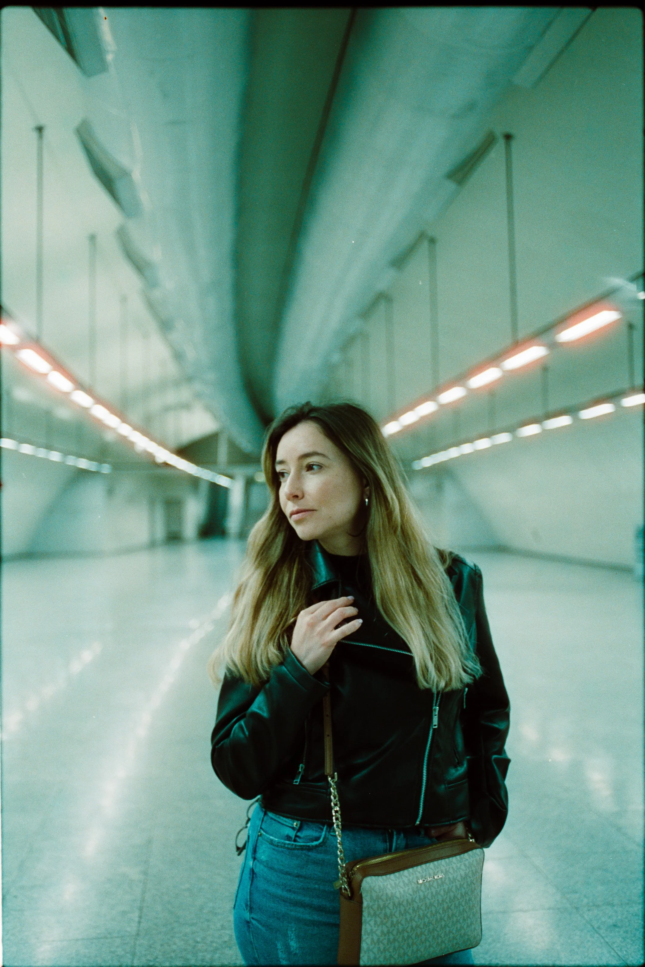 A woman with long blonde hair, wearing a black leather jacket and blue jeans, standing in an empty tunnel, looking to her left with one hand near her neck, holding a brown purse with a chain strap.