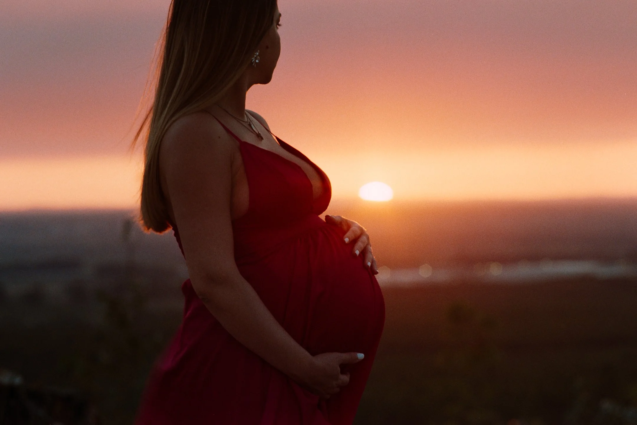 A pregnant woman in a red dress stands by the beach during sunset, cradling her belly with both hands.