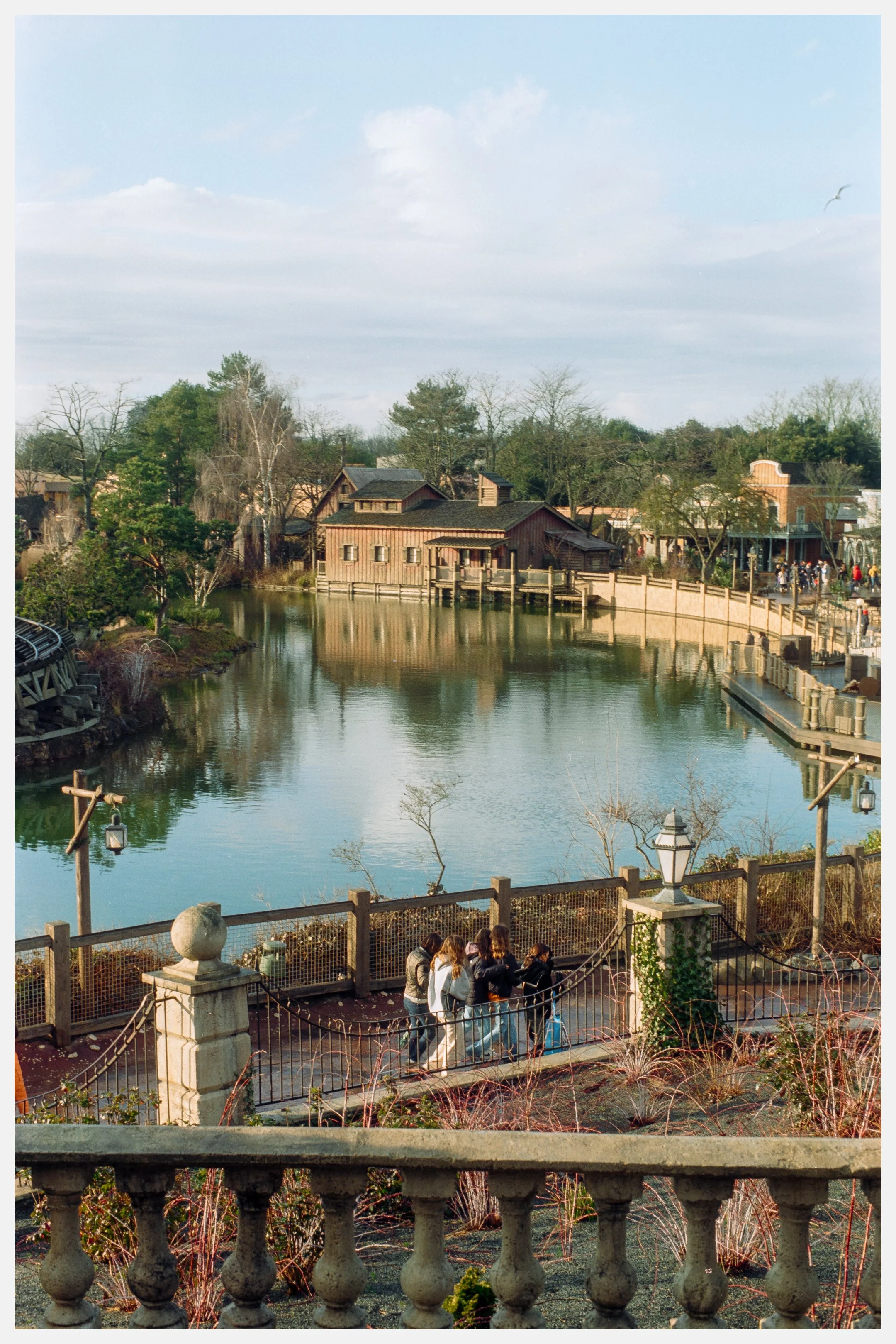 A scenic view of a pond with wooden buildings in a rustic style, surrounded by trees and a walkway, with a group of people walking along a fenced path in the foreground and a bird flying in the sky.
