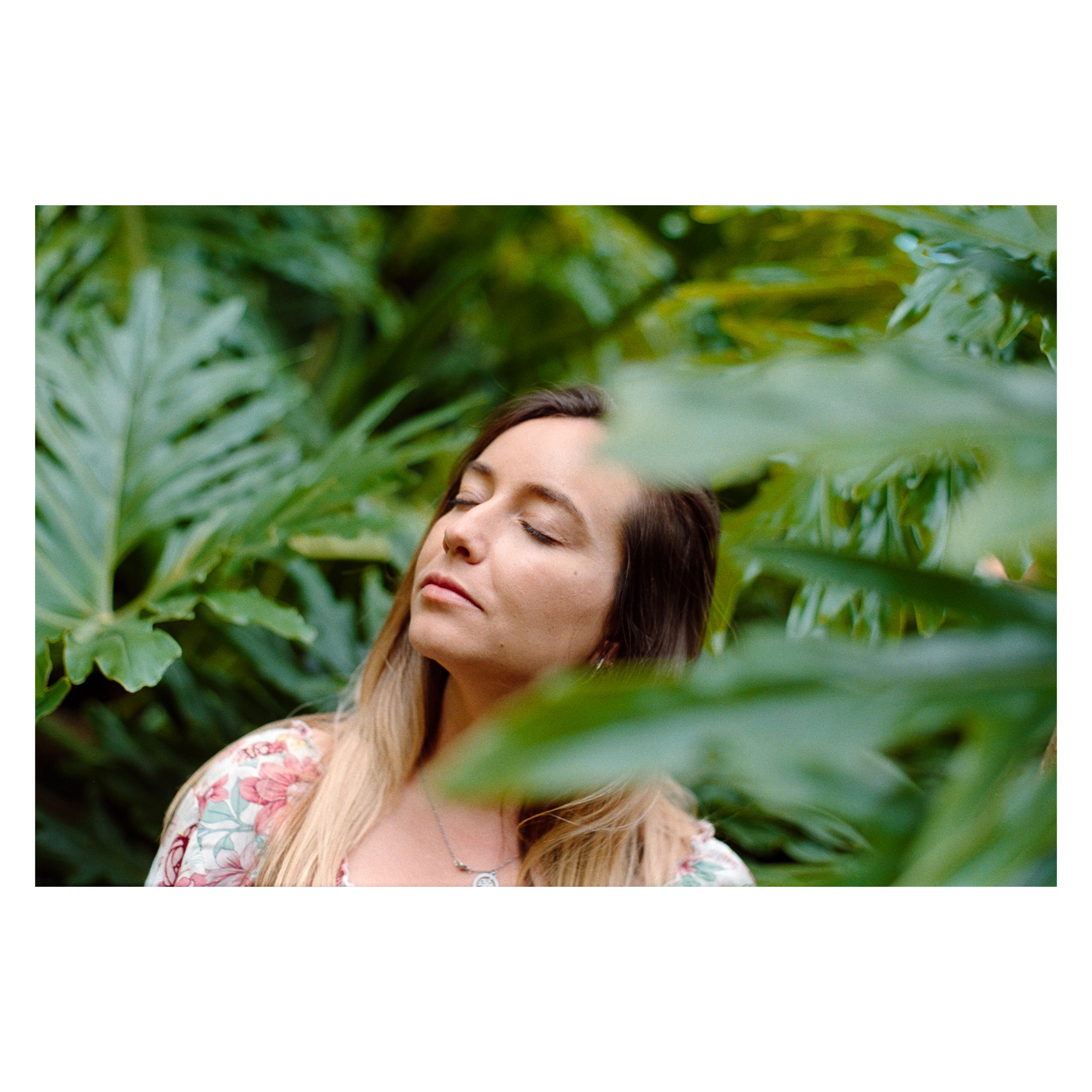 A woman with long brown and blonde hair, wearing a floral pattern top and a silver necklace, has her eyes closed and appears to be enjoying a moment in a lush green jungle filled with large leaves.