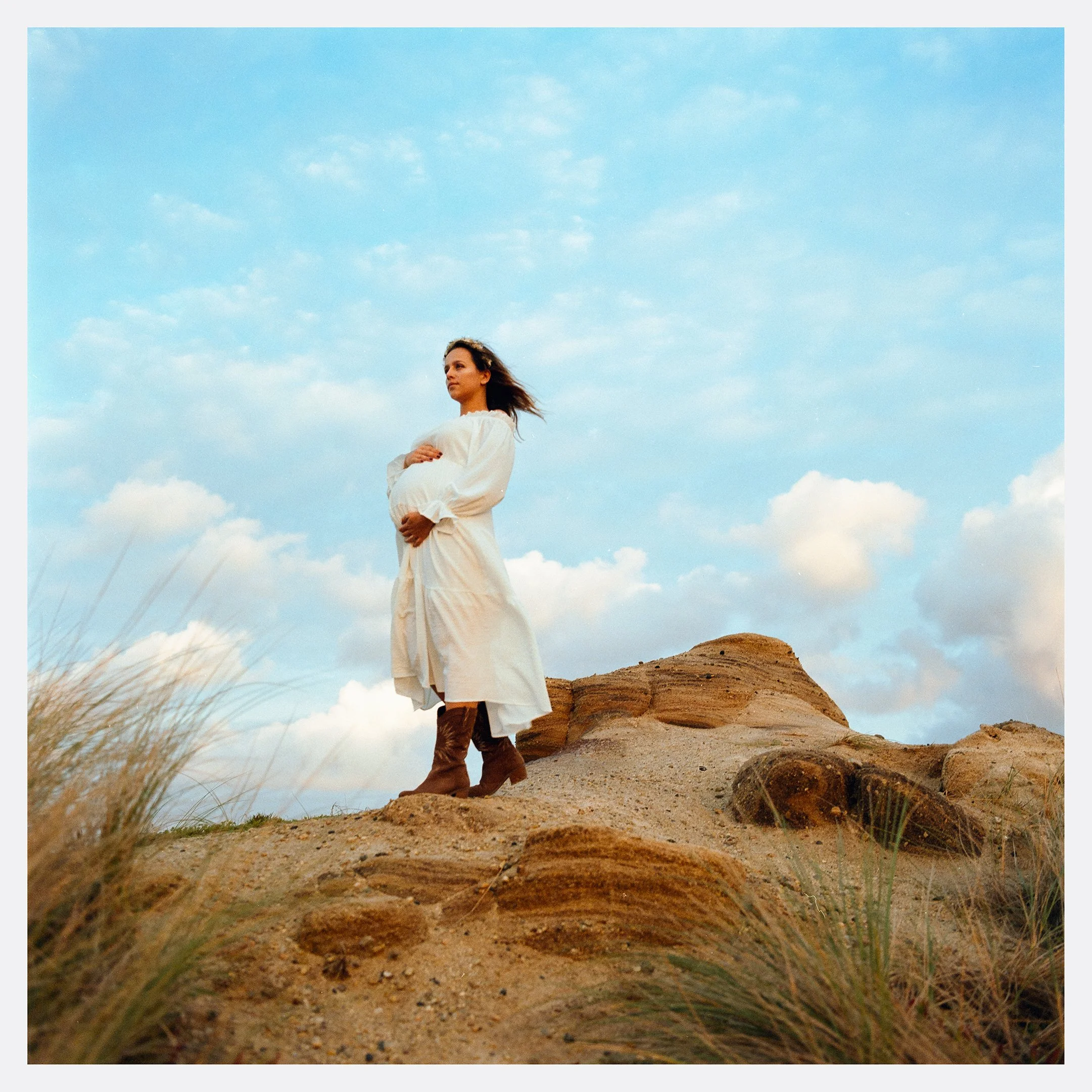A pregnant woman in a white dress standing on a rocky hill with her arms crossed, looking into the distance under a partly cloudy sky.