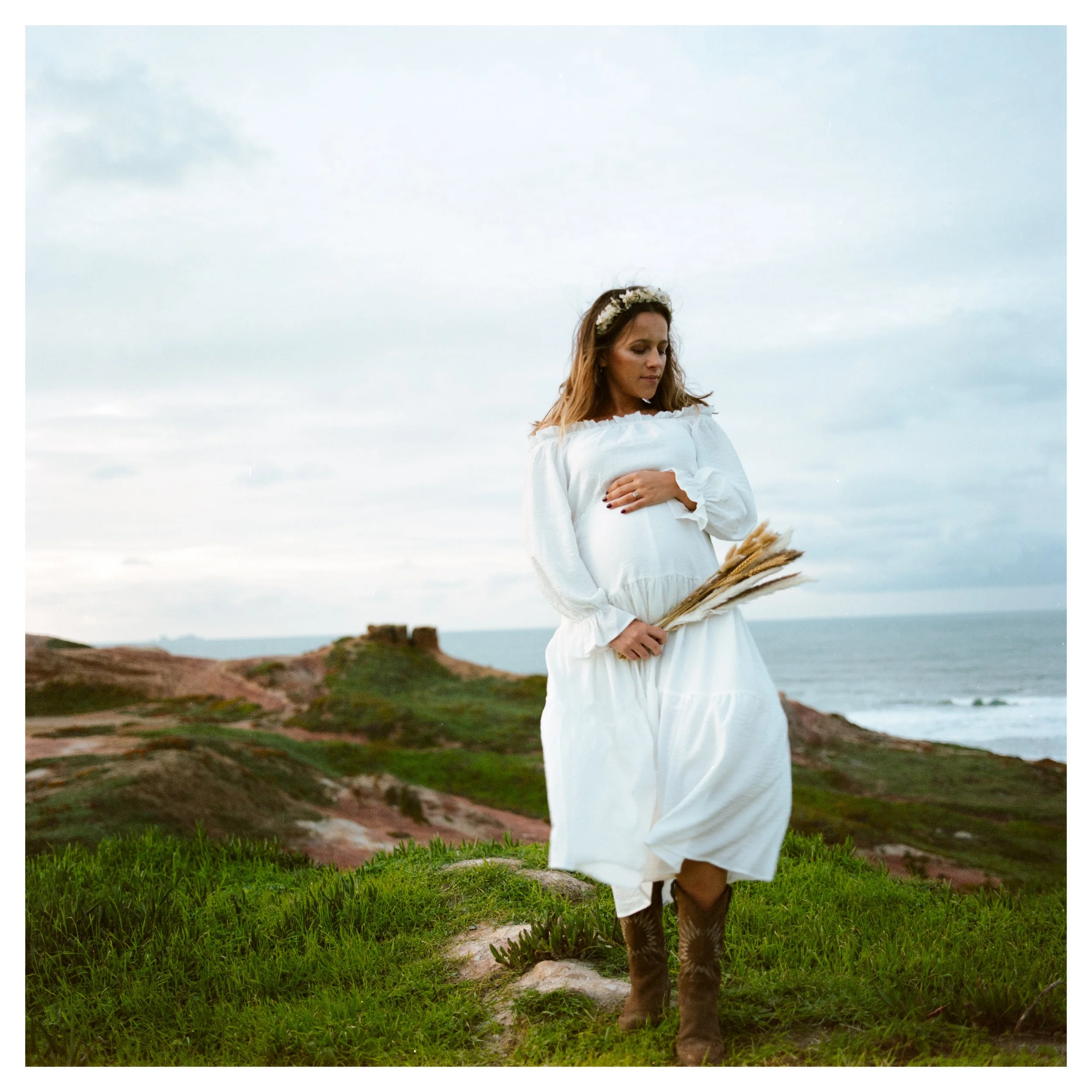 Pregnant woman in white dress holding wheat stalks standing outdoors on grassy hill with ocean and cloudy sky in the background.