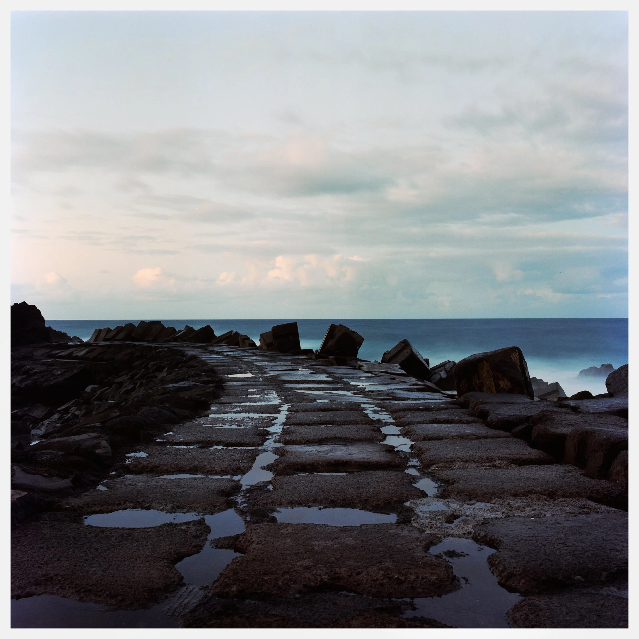 A stone walkway leading towards the ocean with large rocks on either side under a cloudy sky.