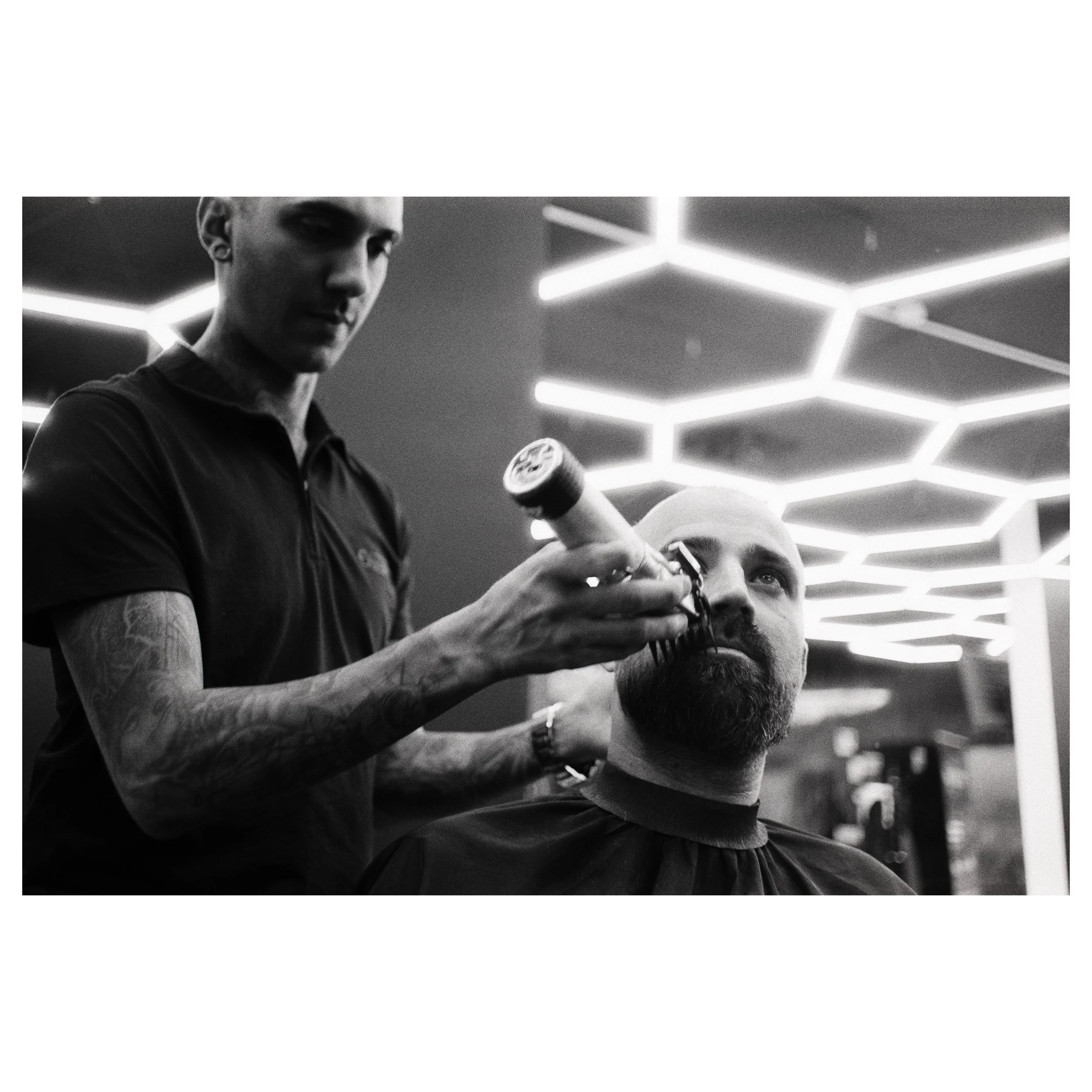 Barber shaving a man's beard with a straight razor in a modern barber shop with geometric ceiling lights. photo by Bruno Novais