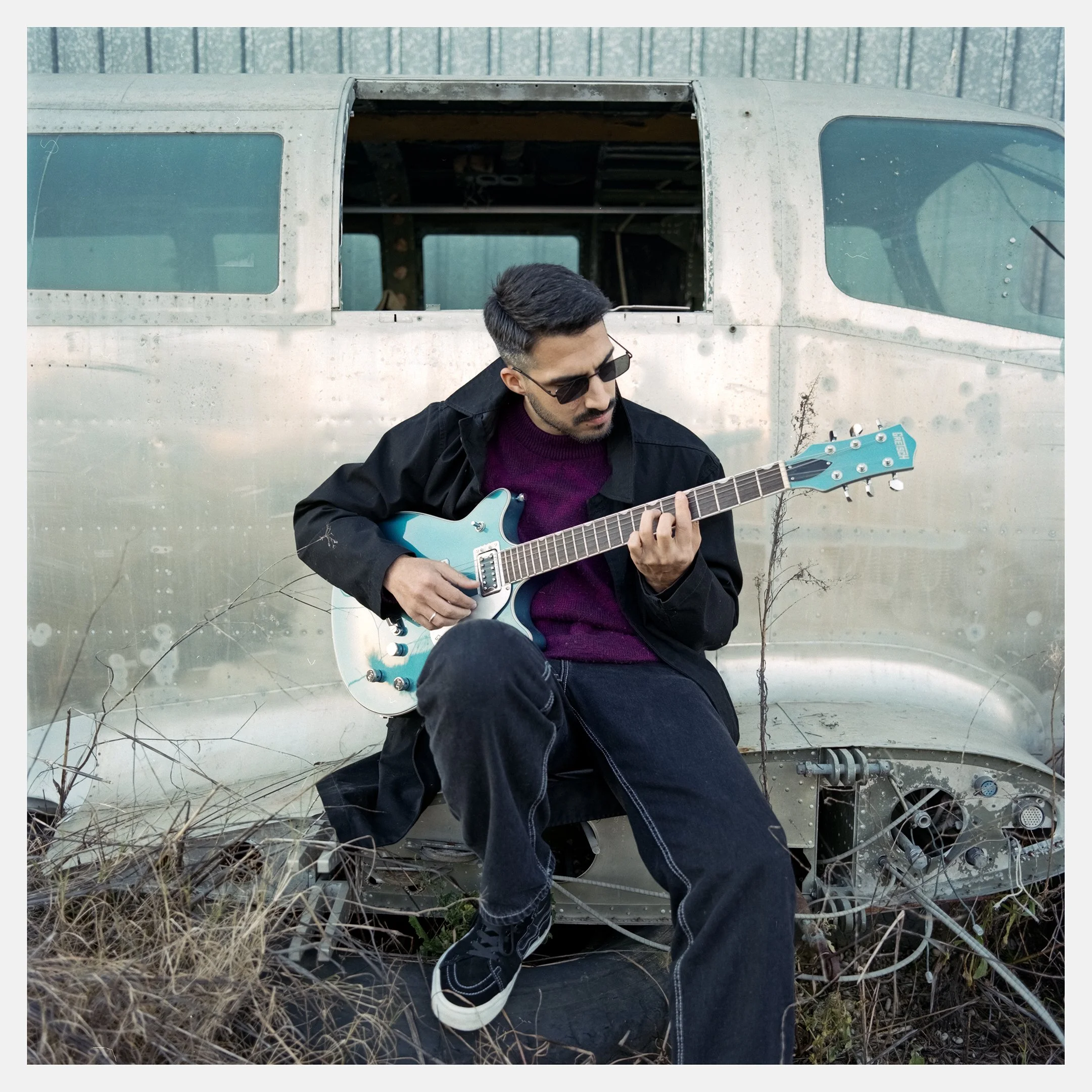 A man wearing sunglasses, a black jacket, and a purple sweater playing an electric guitar while sitting in front of an abandoned, rusty airplane.