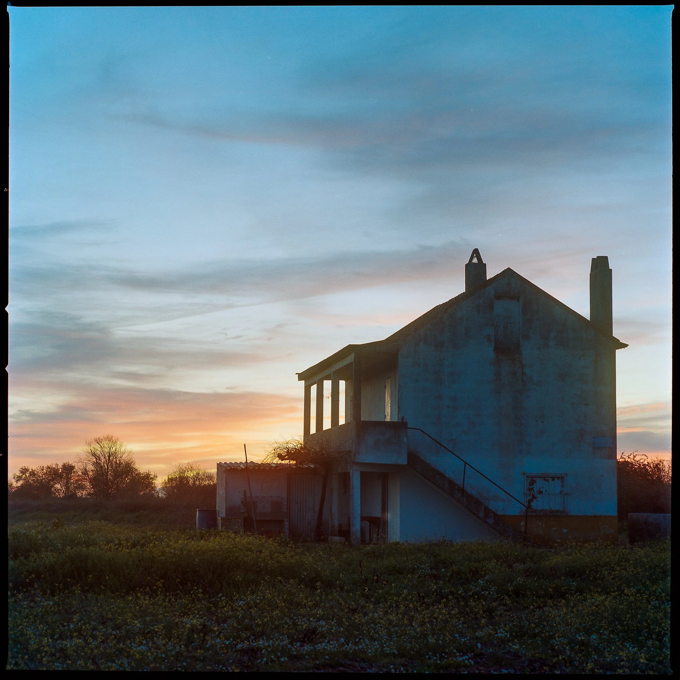 An old, weathered house with a staircase and a small balcony, set against a colorful sunset sky with clouds and trees in the background.