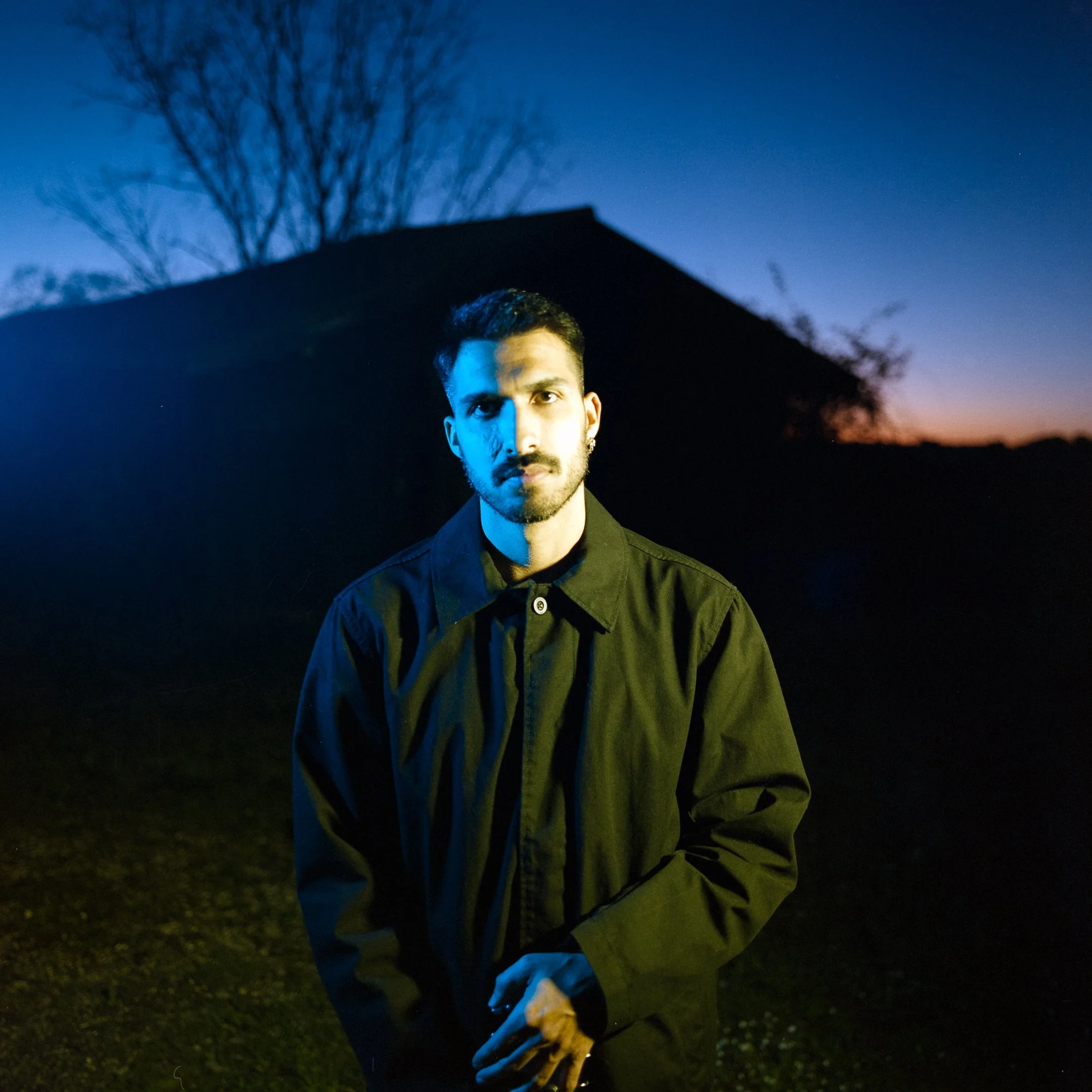 A man with dark hair, a beard, and earrings stands outdoors at dusk, illuminated by blue and yellow lighting, with a dark silhouette of a house and leafless trees in the background.