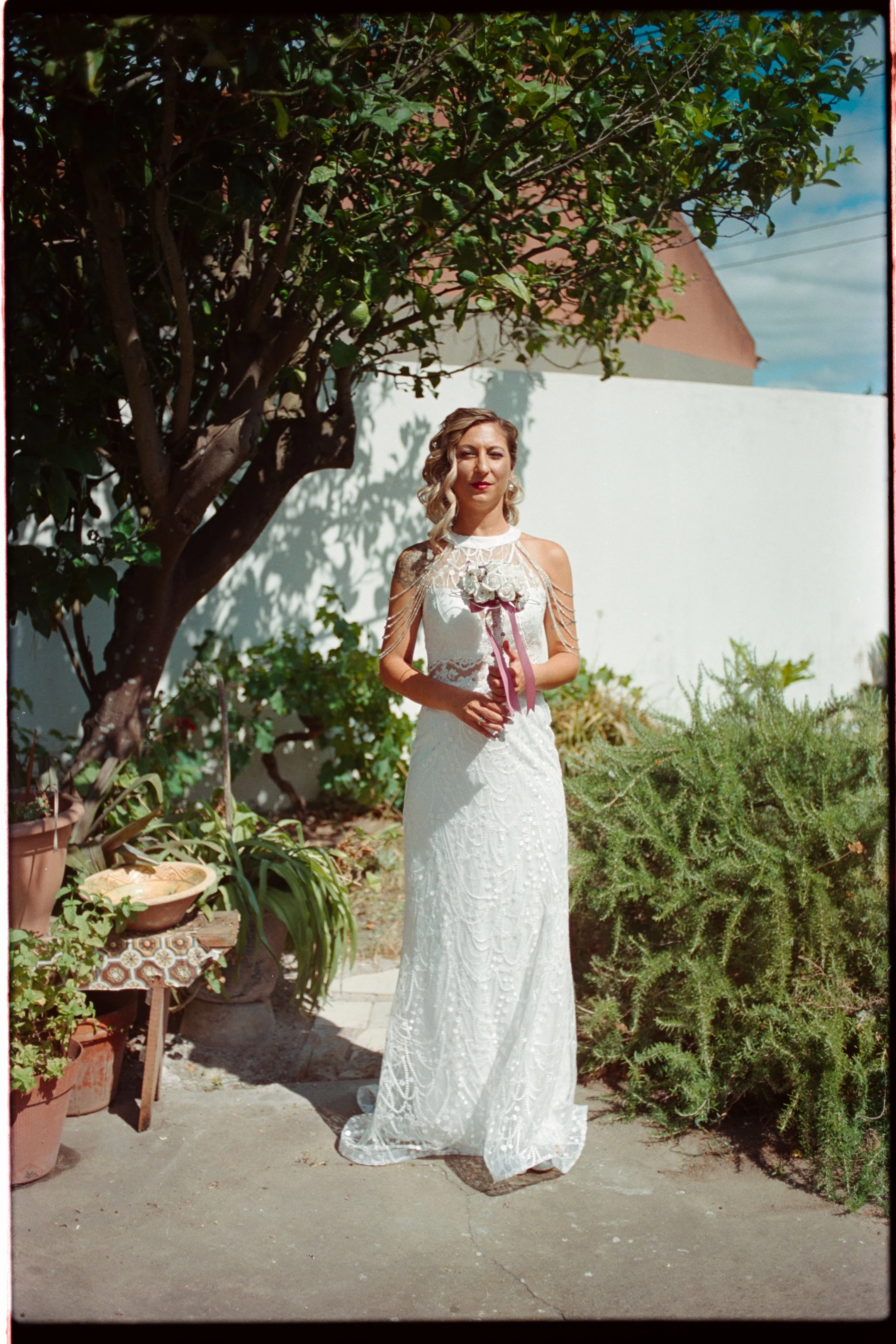A woman in a white bridal gown holding a bouquet standing outdoors in a garden with plants and trees under a blue sky. photo by Bruno Novais