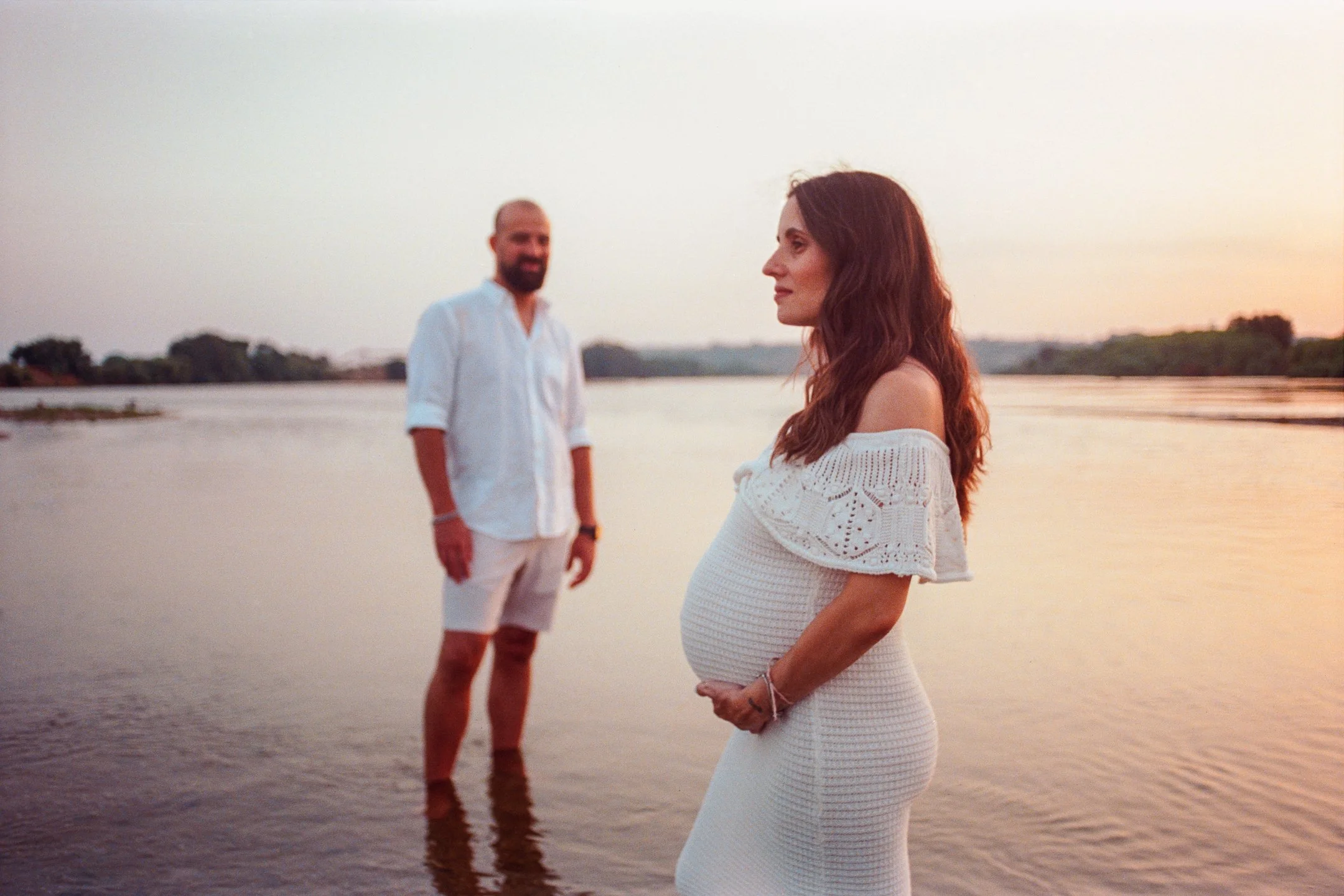 A pregnant woman in a white dress standing by a river at sunset, with a man in a white shirt and shorts standing in the water in the background. photo by Bruno Novais