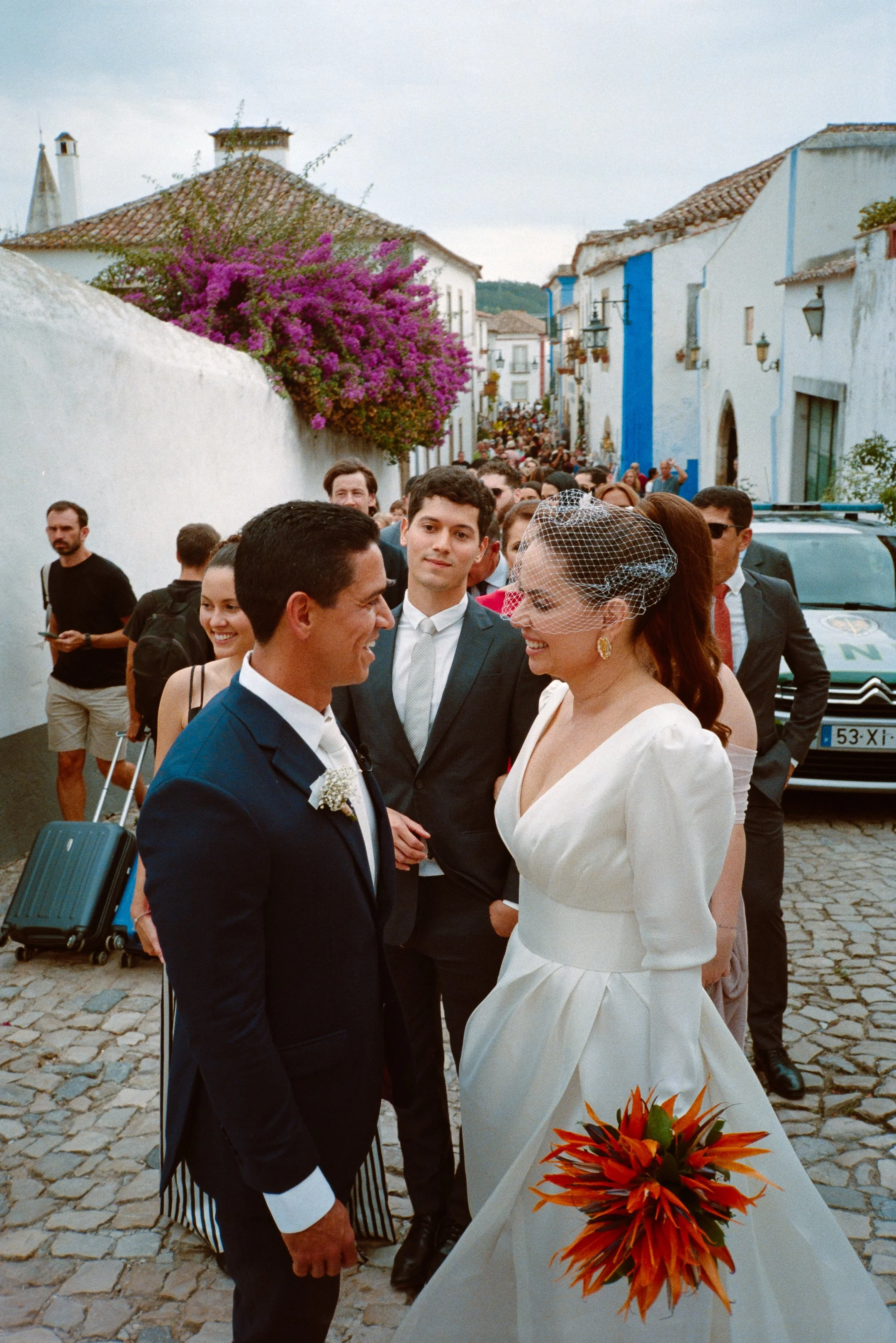 A wedding scene on a cobblestone street with a bride in a white dress with a veil, holding a bouquet of orange flowers, talking to a groom in a dark suit with a white boutonniere, surrounded by friends and family.photo by Bruno Novais