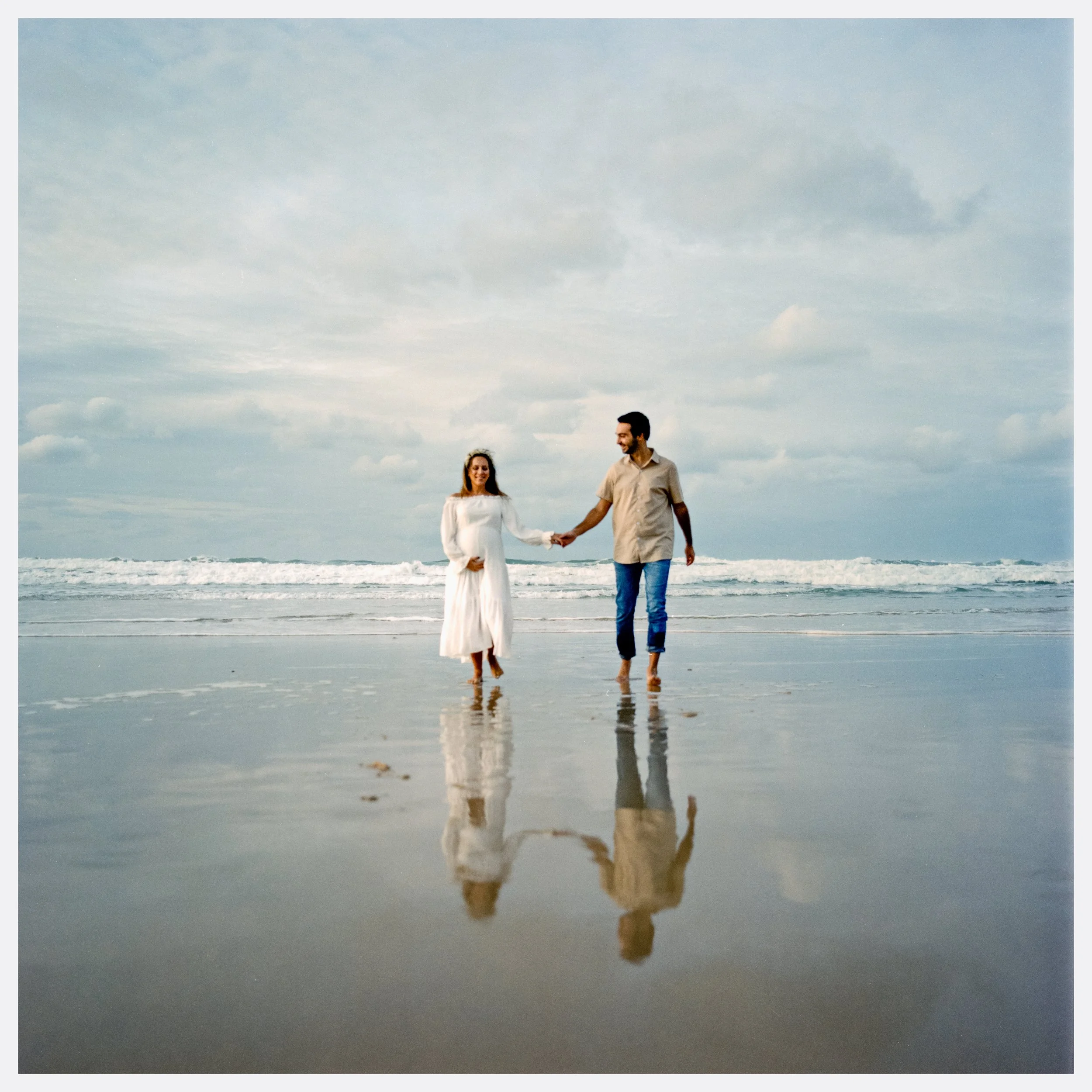 A couple holding hands, walking along the beach at sunset, with their reflections visible in the shallow water.