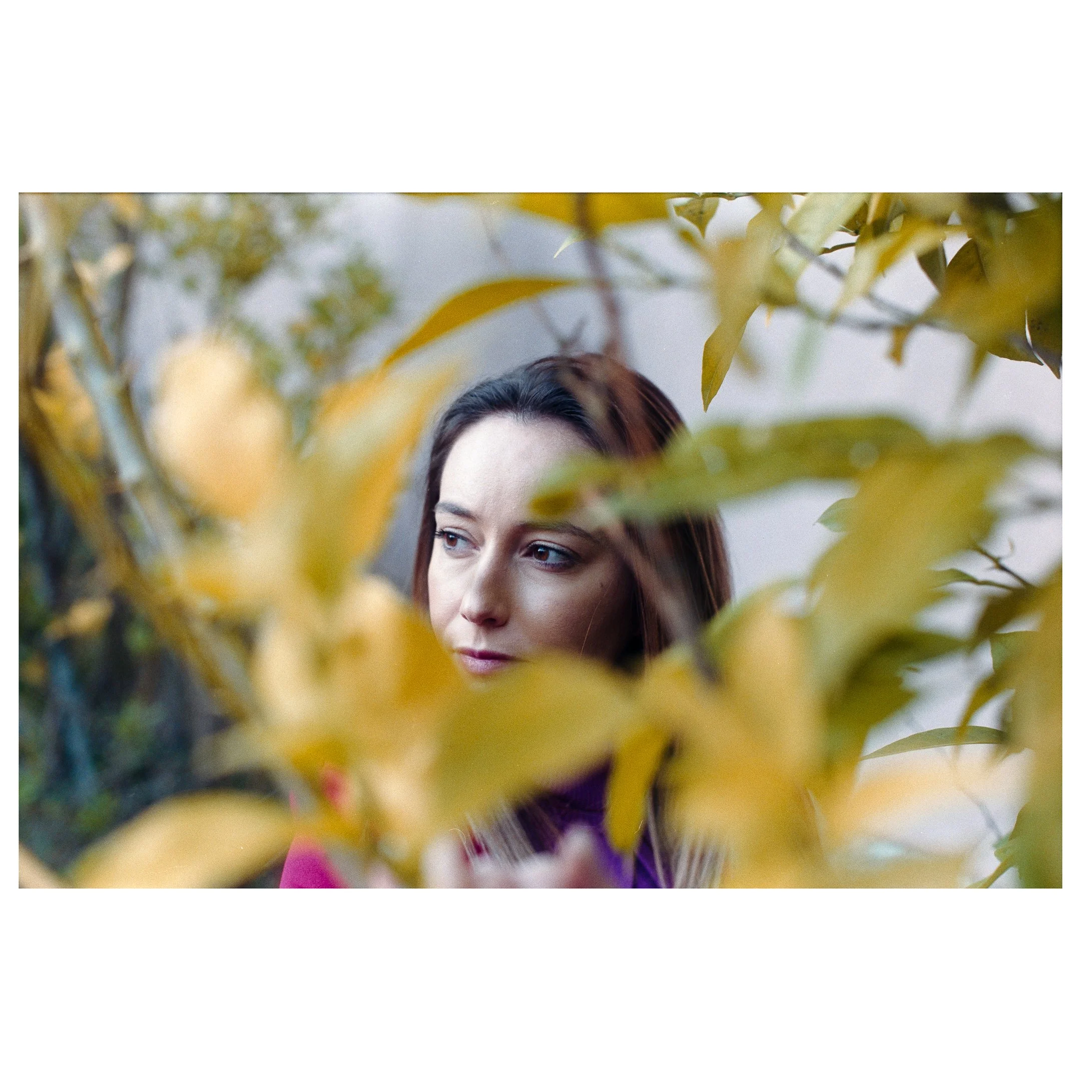 Young woman with brown hair and light skin looking through yellow and green leaves.