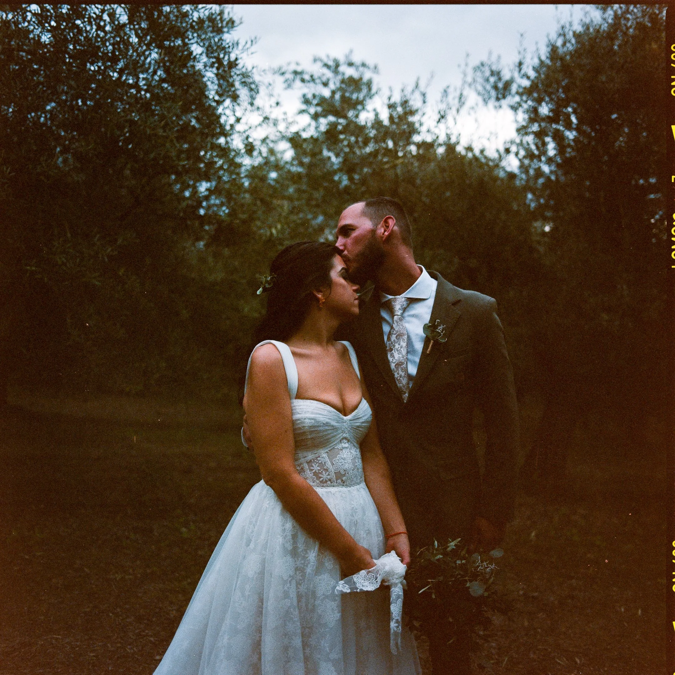 A newlywed couple standing outdoors, the groom kissing the bride on the forehead, with trees and a cloudy sky in the background.
