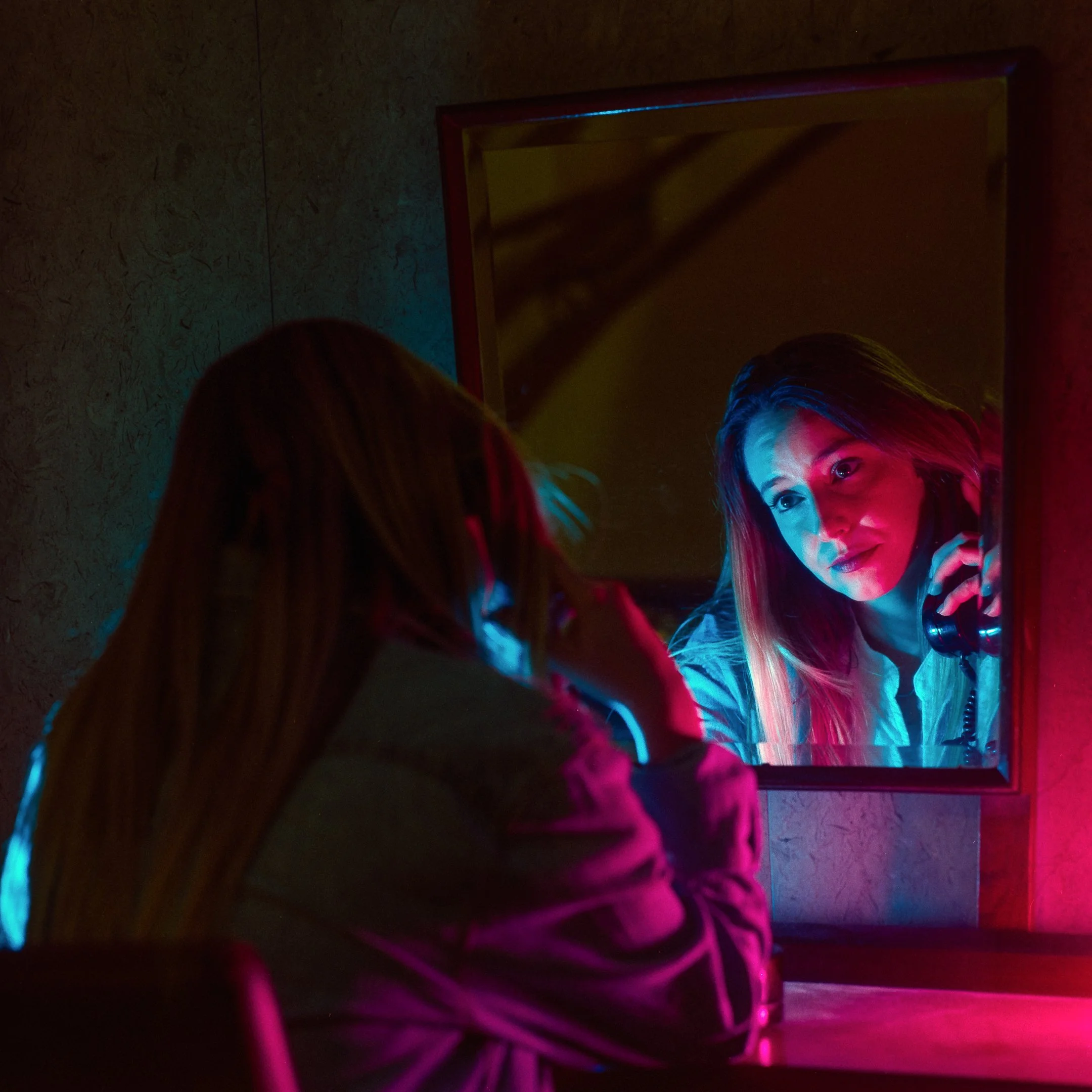 A woman with long hair looking at her reflection in a mirror, illuminated by colorful pink and blue neon lights, holding a phone to her ear.