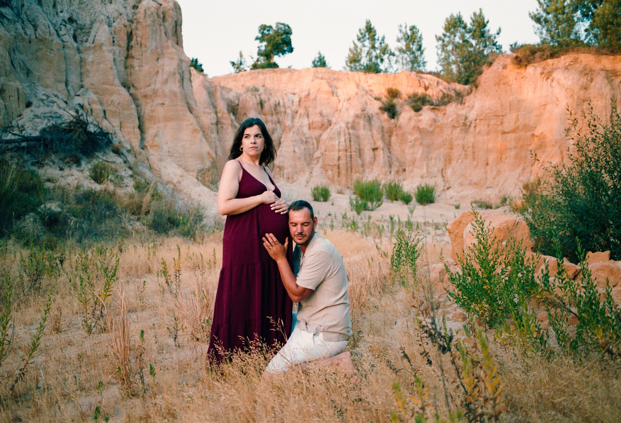A pregnant woman in a maroon dress standing outdoors in a desert landscape with a man kneeling beside her, holding her hand and resting his head on her belly.
