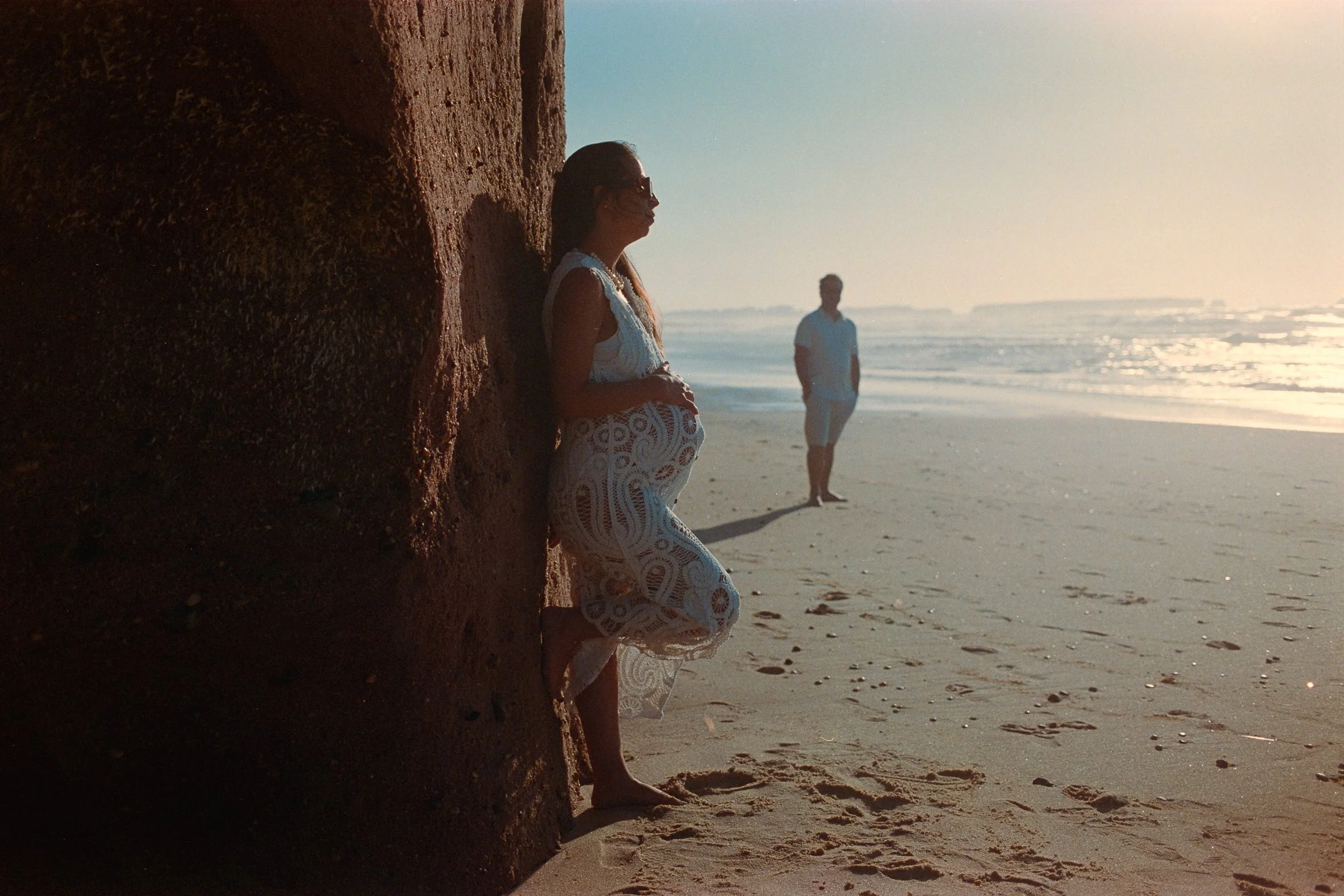 A pregnant woman in a white lace dress leaning against a large rock on the beach, with a man walking in the background near the shoreline during sunset.