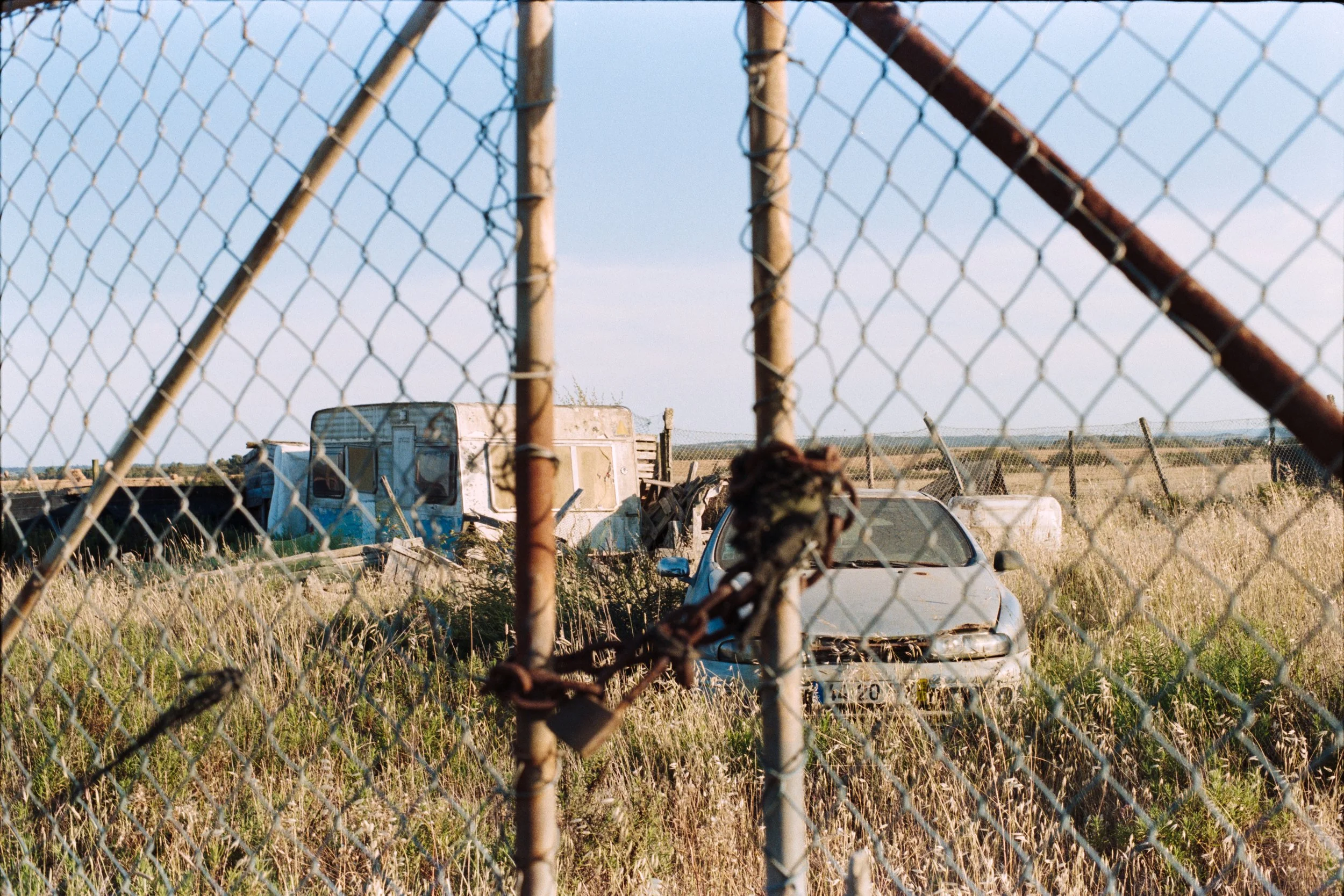 An old, abandoned car and a dilapidated camper behind a rusty chain-link fence in a grassy field, with a distant horizon and cloudy sky.