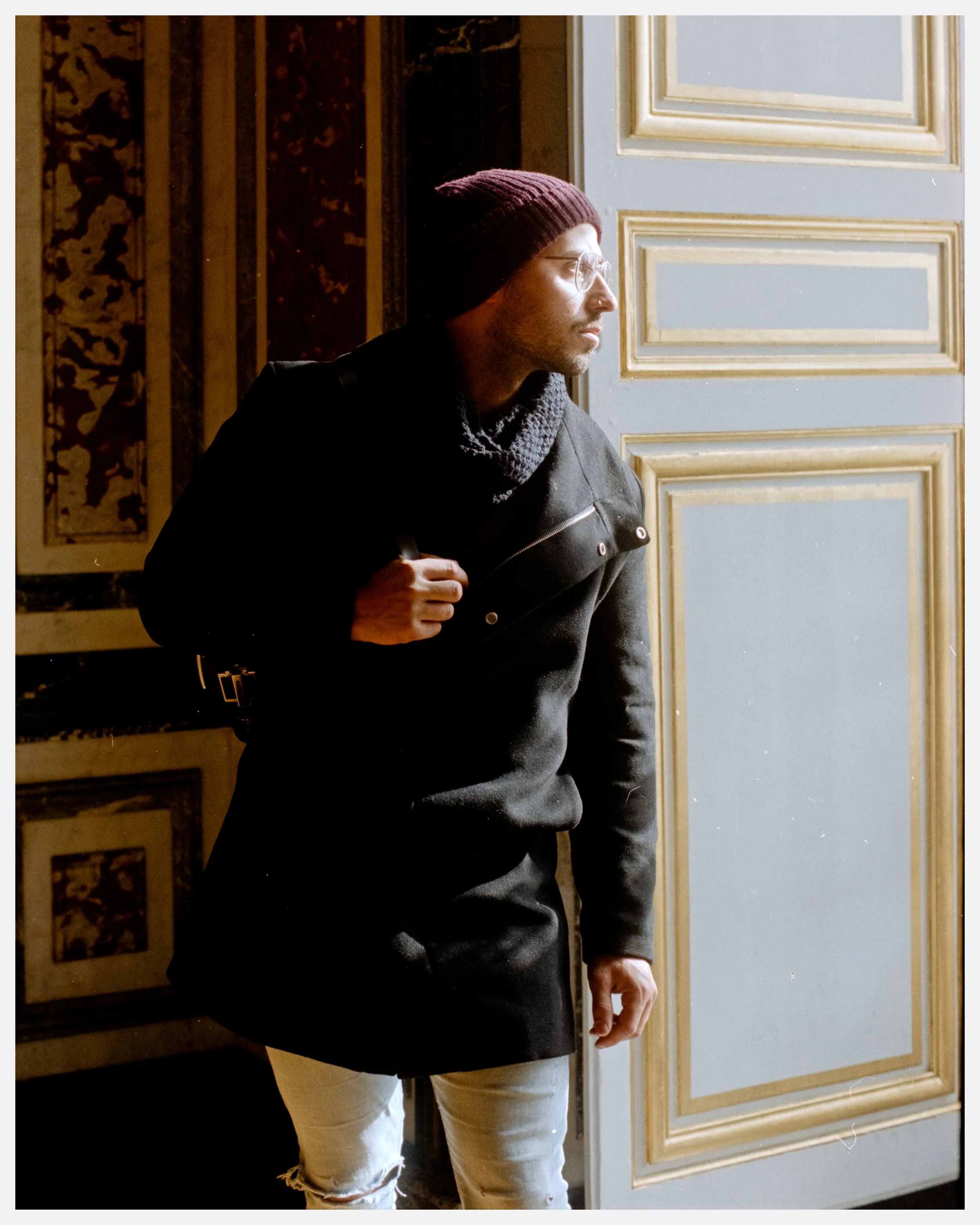 A man in a black jacket, knit hat, and scarf gazes out of a window in an ornate interior with gold accents.