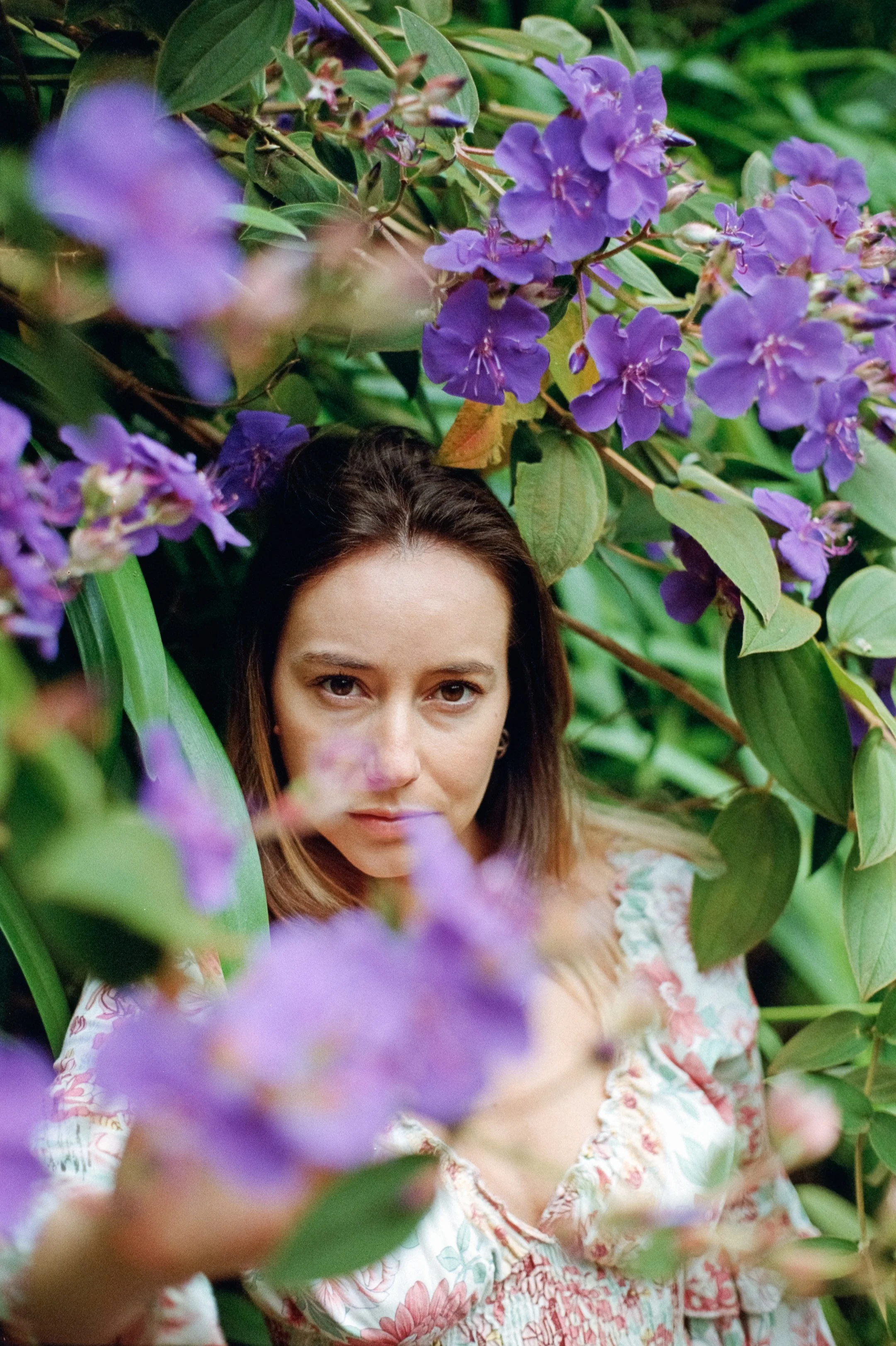 A woman with light skin and brown hair is surrounded by lush green foliage and purple flowers, gazing into the camera with a neutral expression.