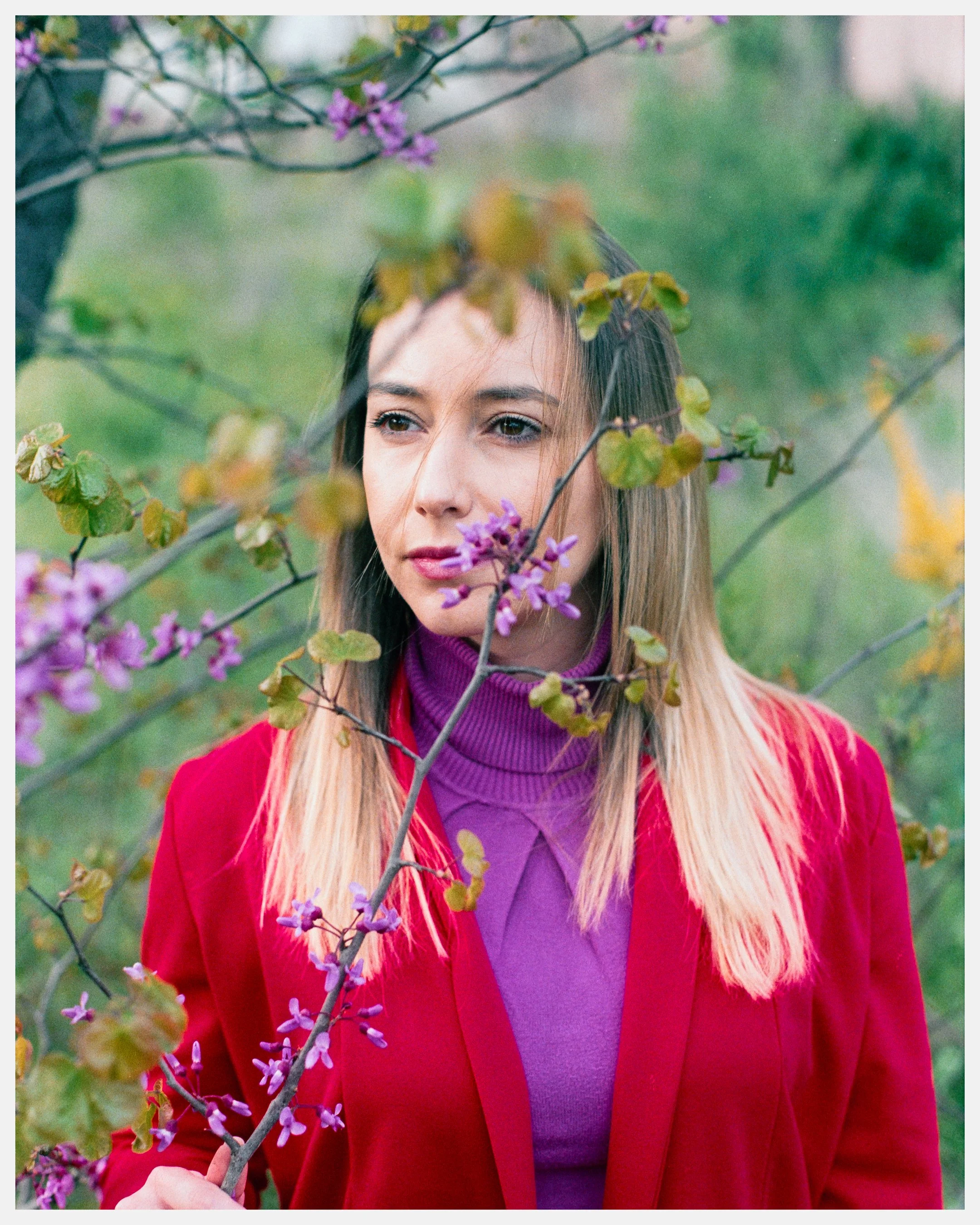 A woman with long blonde hair standing outdoors among flowering branches. She wears a purple turtleneck and a red blazer, gazing thoughtfully into the distance.