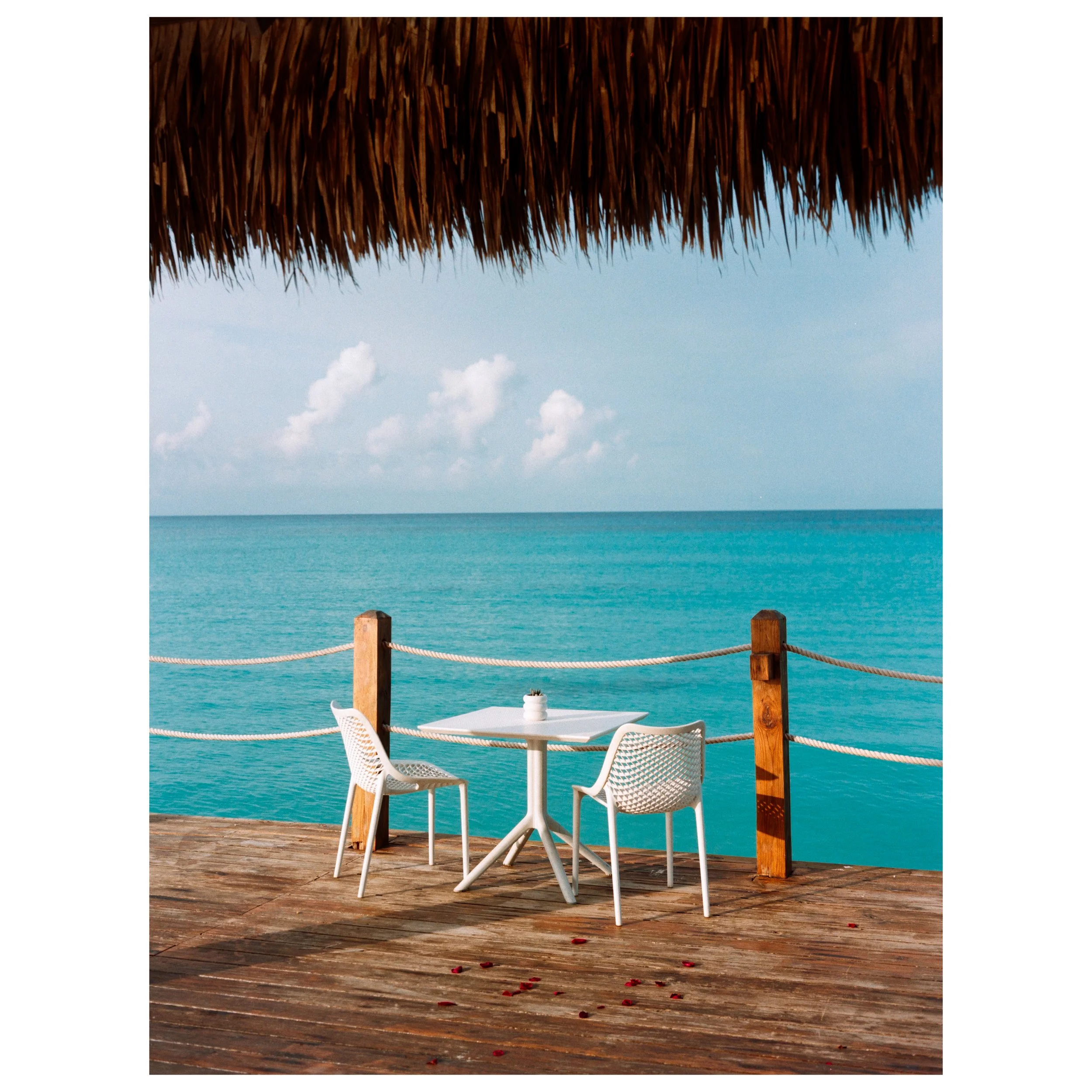 A wooden deck with an empty white table and two white chairs overlooking the ocean. The scene is shaded by a thatched roof, with a rope fence along the edge of the deck. The sky is partly cloudy with calm turquoise water in the background.