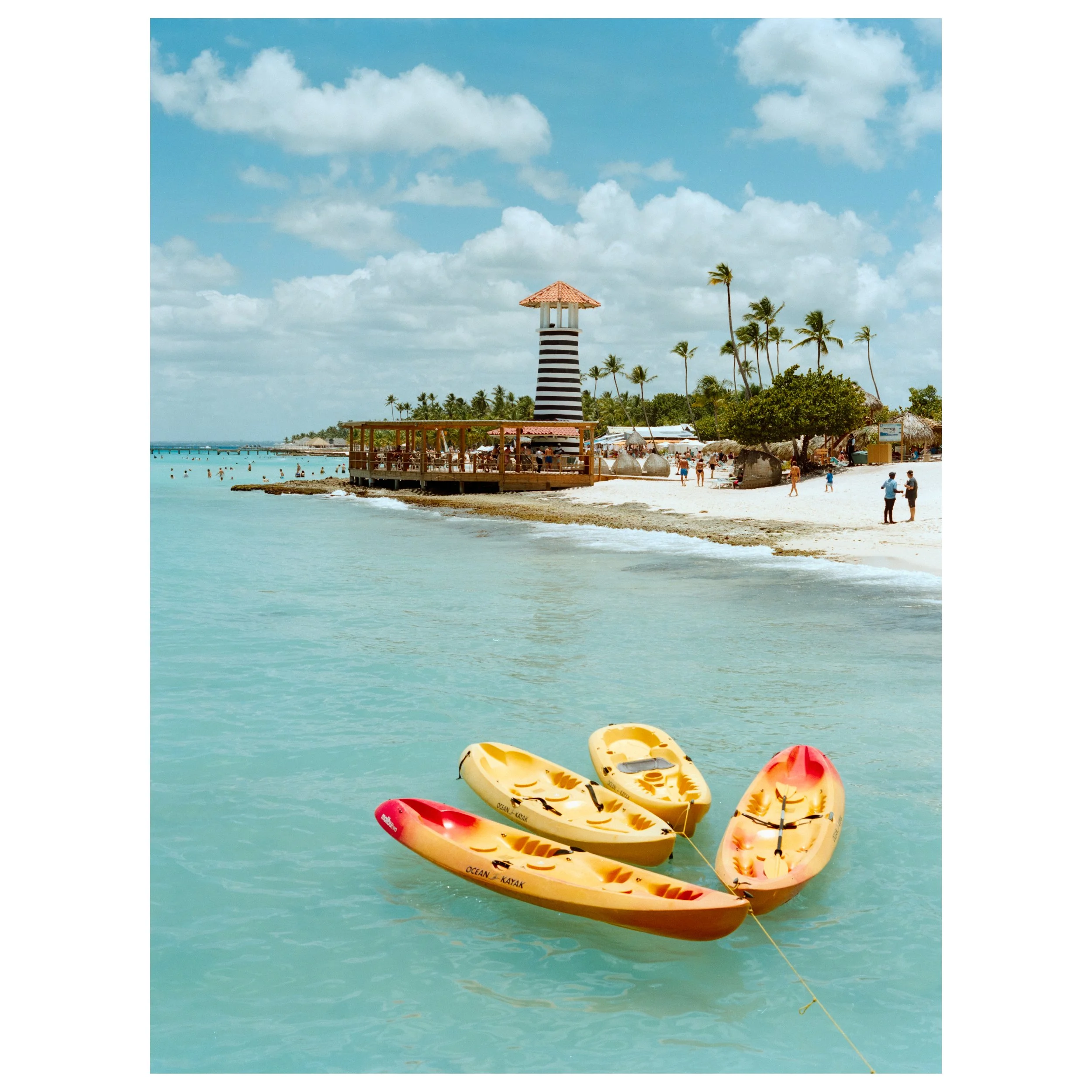 Colorful kayaks floating in turquoise water near a sandy beach with palm trees and a lighthouse in the background under a blue sky with clouds.