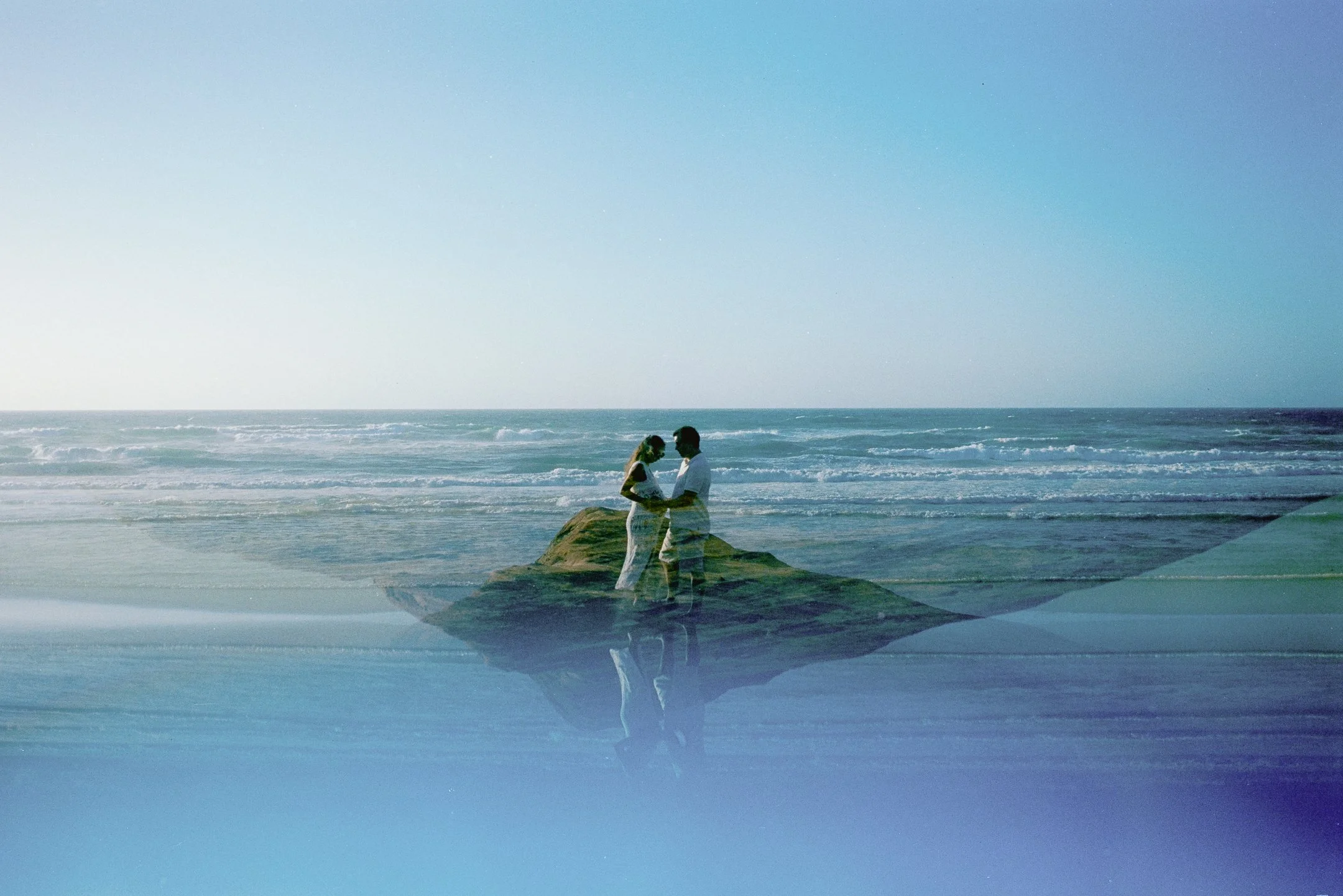 A couple standing on a large rock at the beach, with the ocean and sky in the background, during daytime.