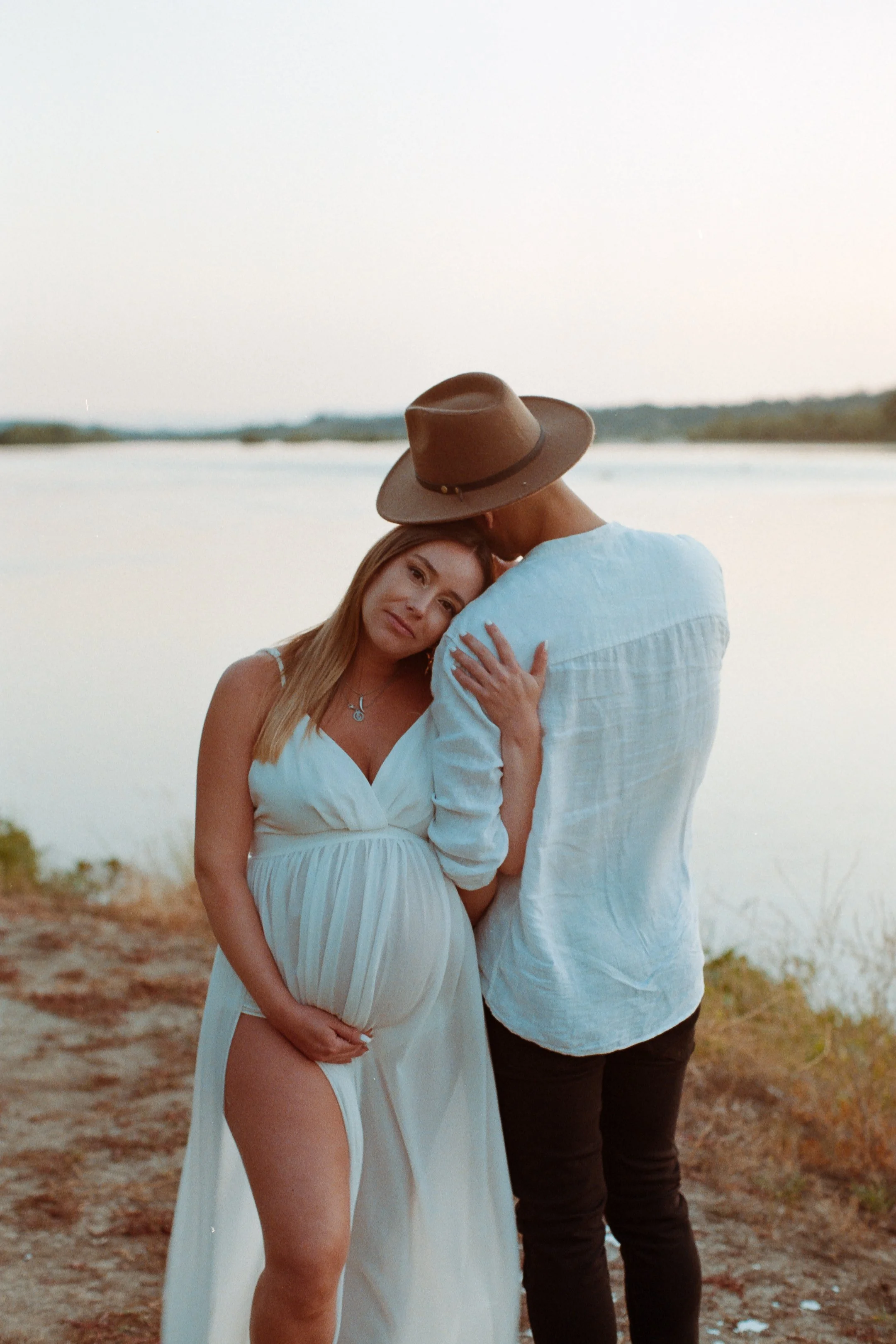 A pregnant woman in a white dress embraces a man wearing a white shirt and a wide-brimmed brown hat, near a body of water at sunset.