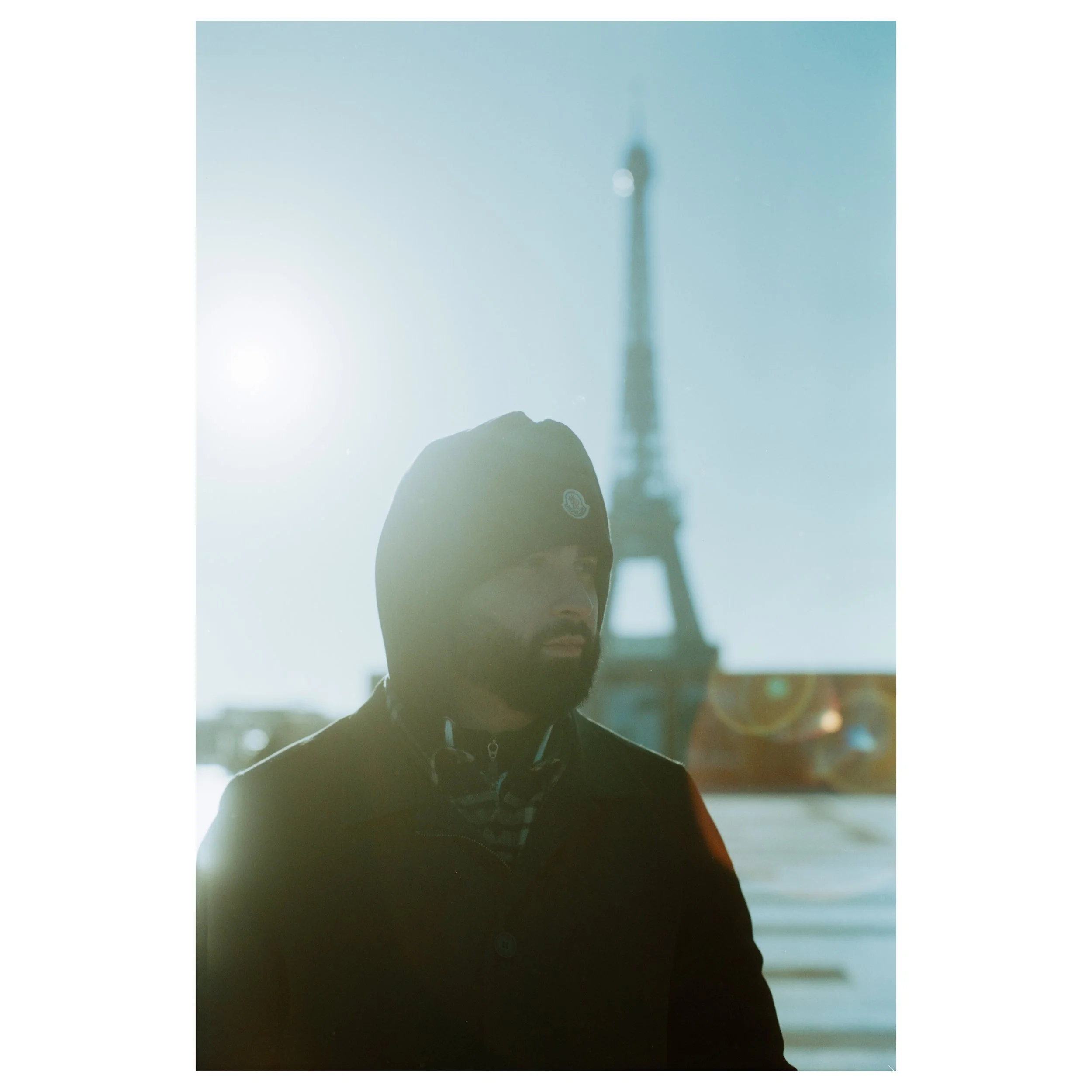 A man wearing a black beanie and jacket stands near a window with sunlight behind him. The Eiffel Tower is visible in the background through the glass.