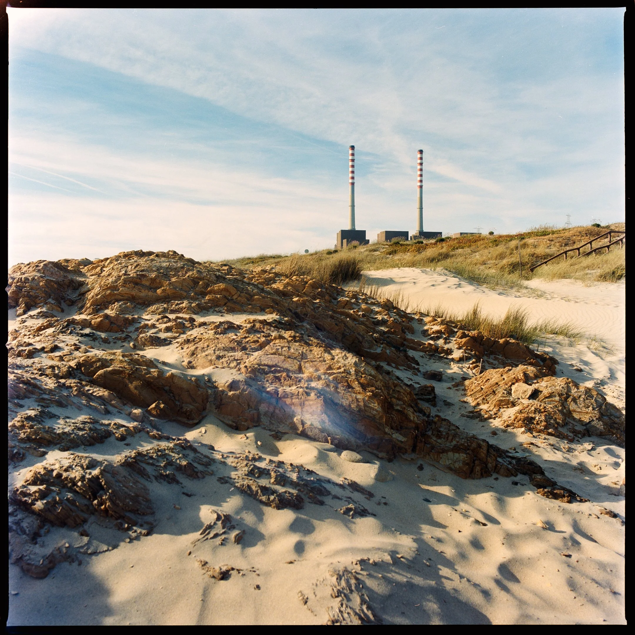 Sand dunes with rocks and grass in the foreground, power plant with two tall smokestacks with red and white stripes in the background under a partly cloudy sky.