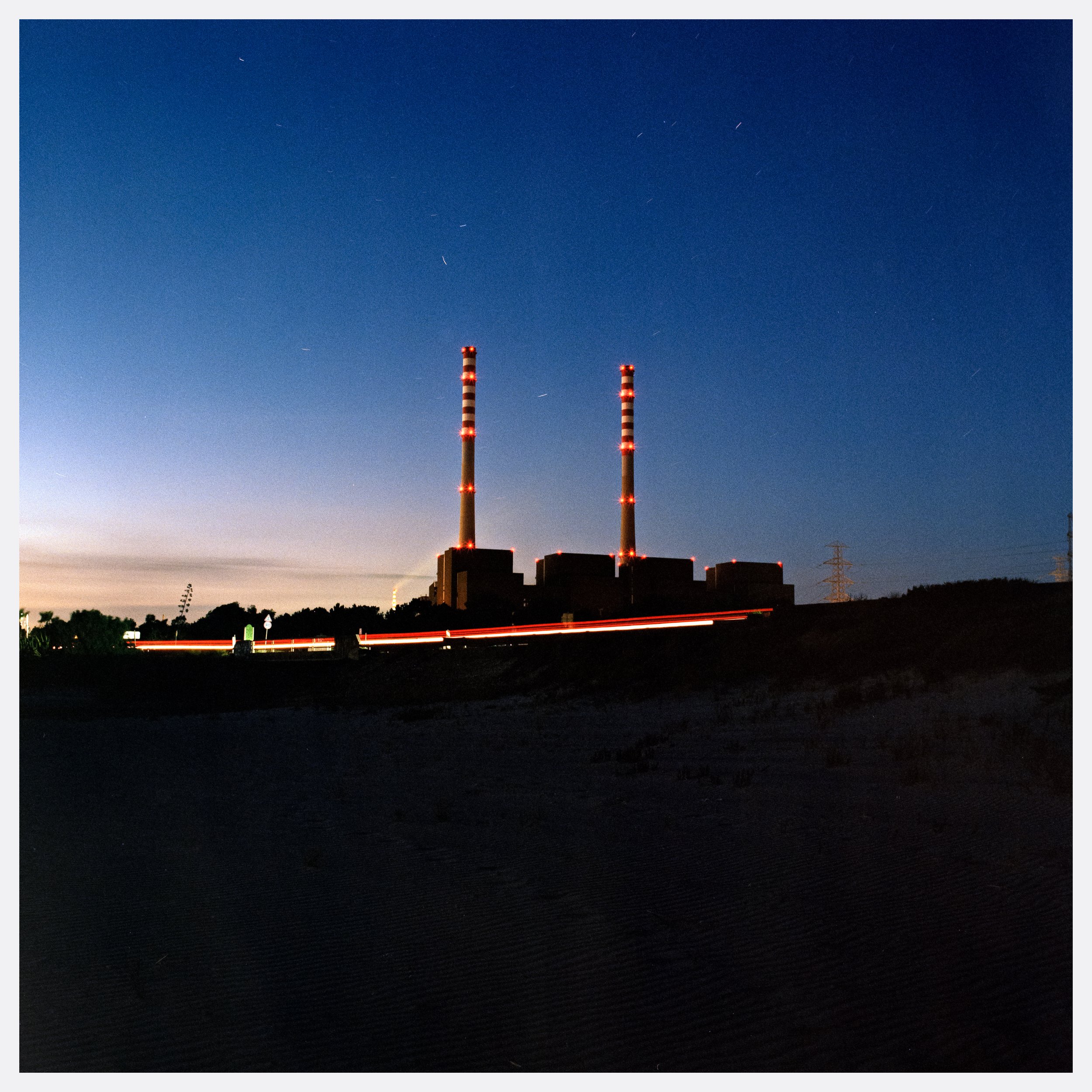 Nighttime industrial scene with two tall smokestacks with red and white lights, dark silhouettes of factory structures, and streaks of light from moving vehicles.
