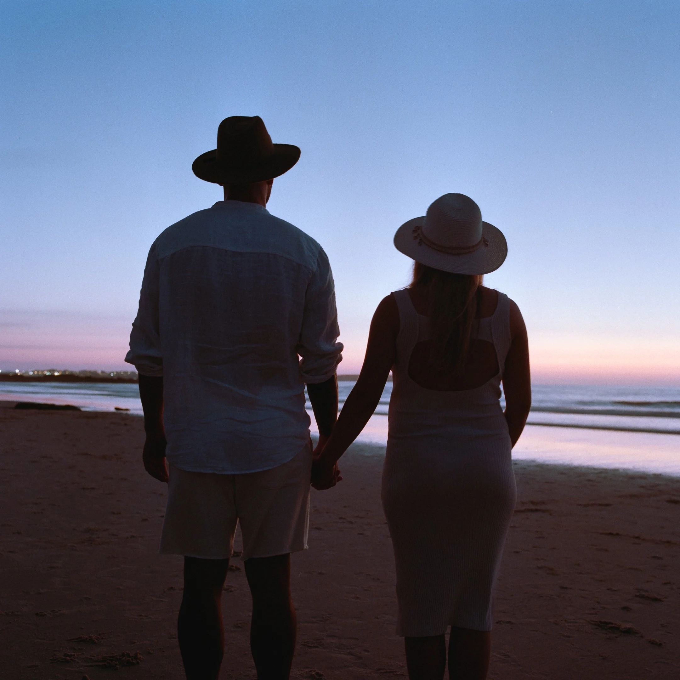 Couple holding hands on the beach at sunset, facing the ocean photo by Bruno Novais