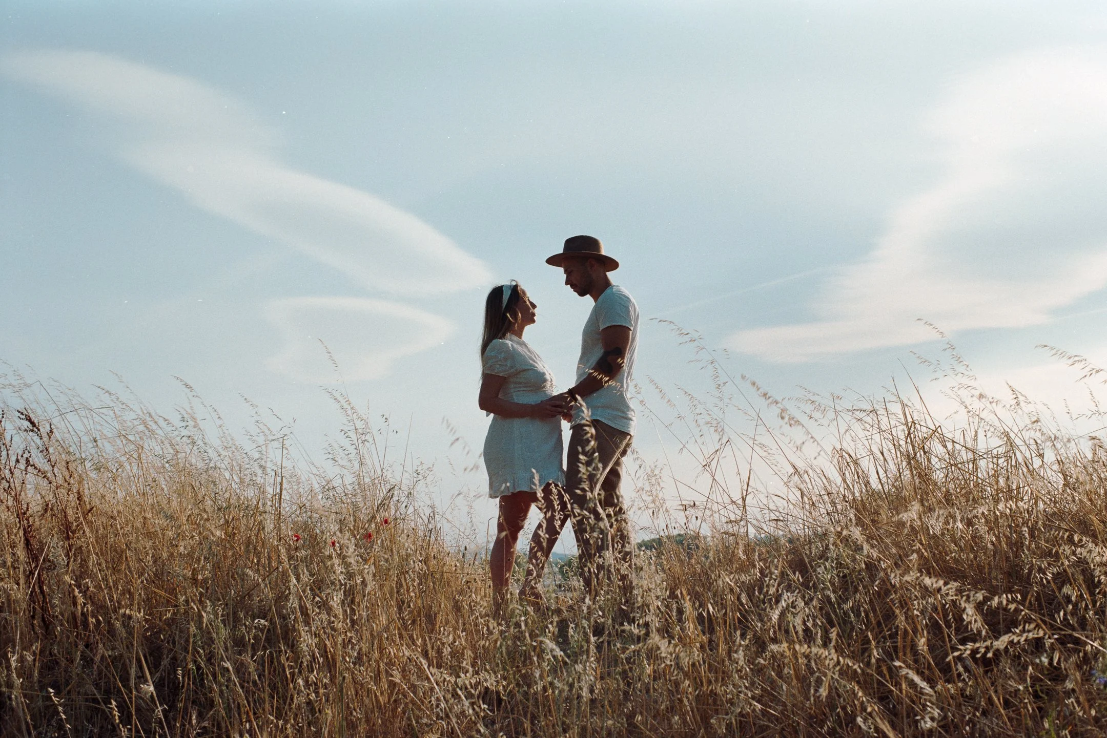 A couple stands close together in a field of tall dry grass, looking at each other, with the man wearing a hat and the woman wearing sunglasses. The sky is mostly clear with a few clouds.