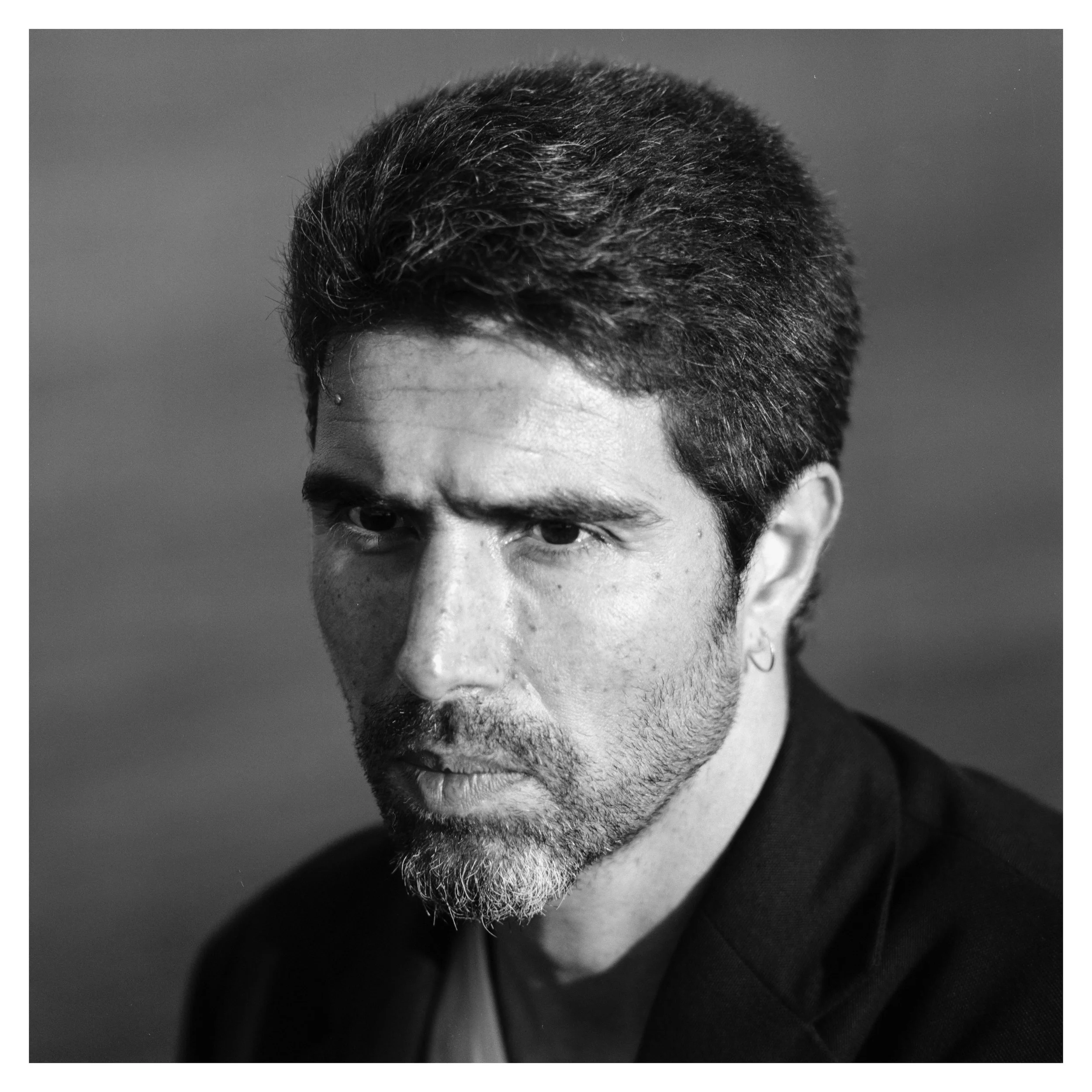 Black and white close-up portrait of a man with dark hair, a beard, and an earring, looking intensely at the camera.