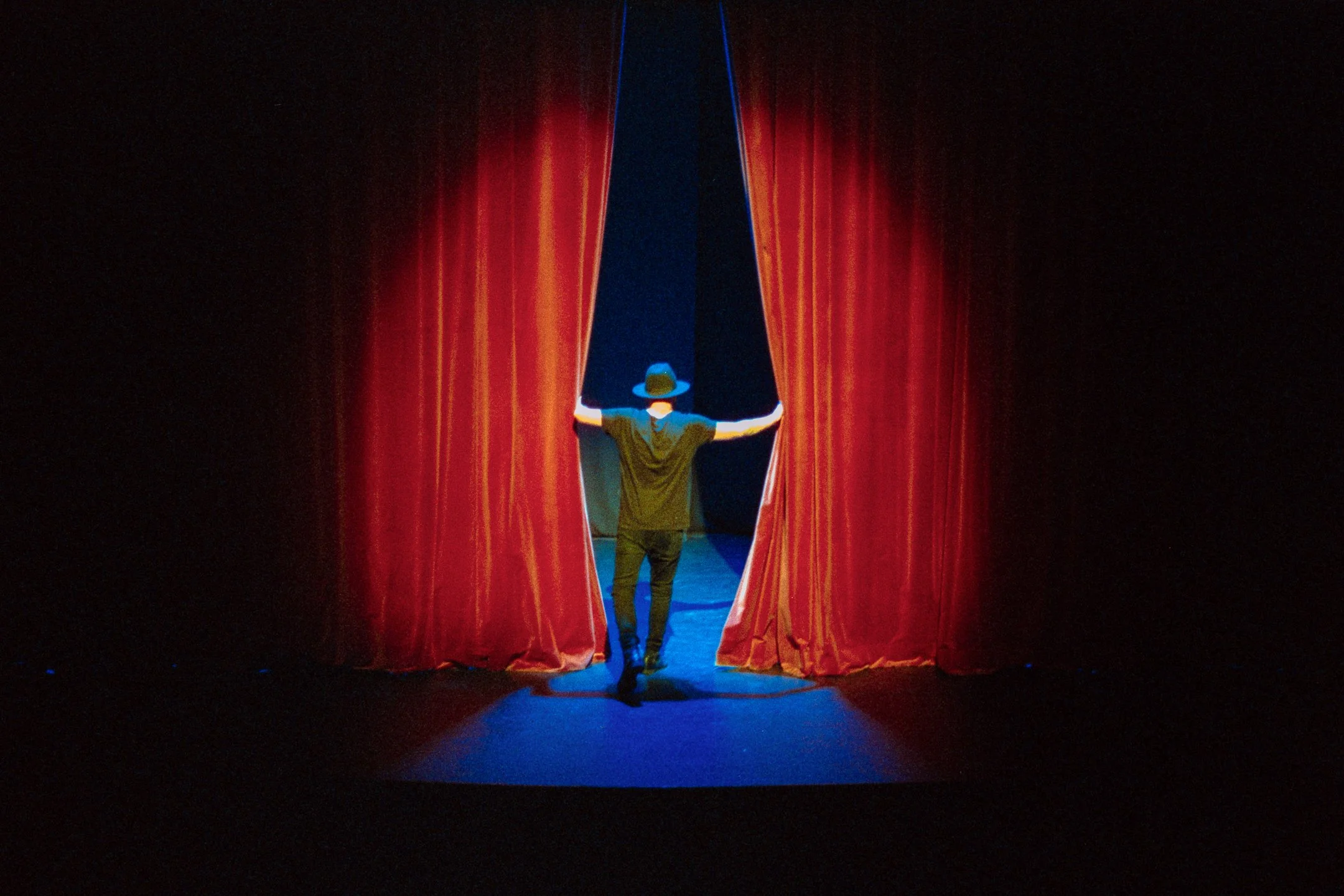 A person wearing a hat and black clothing stands on stage, opening red curtains to reveal a dark background.