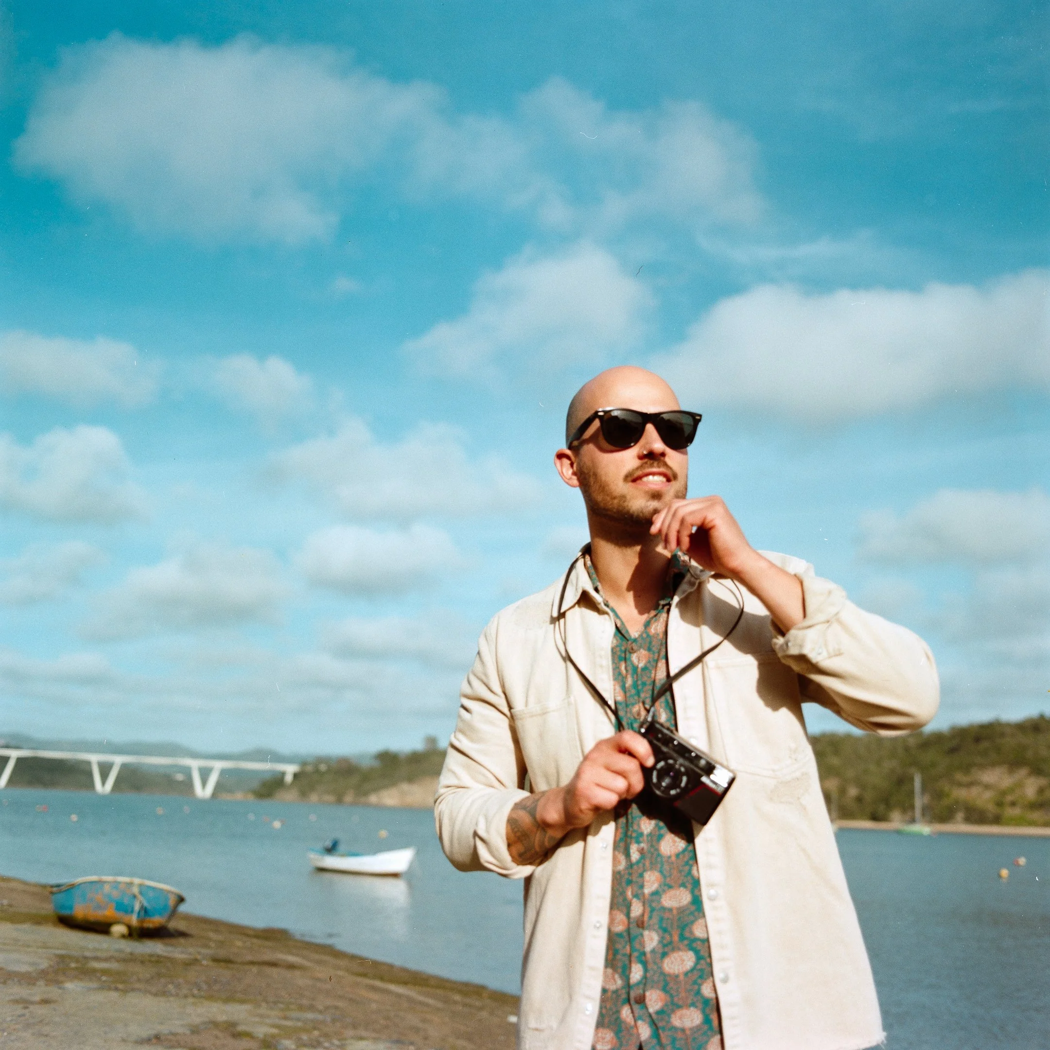 A man with sunglasses holding a camera, standing near a body of water with boats and a bridge in the background under a partly cloudy sky.
