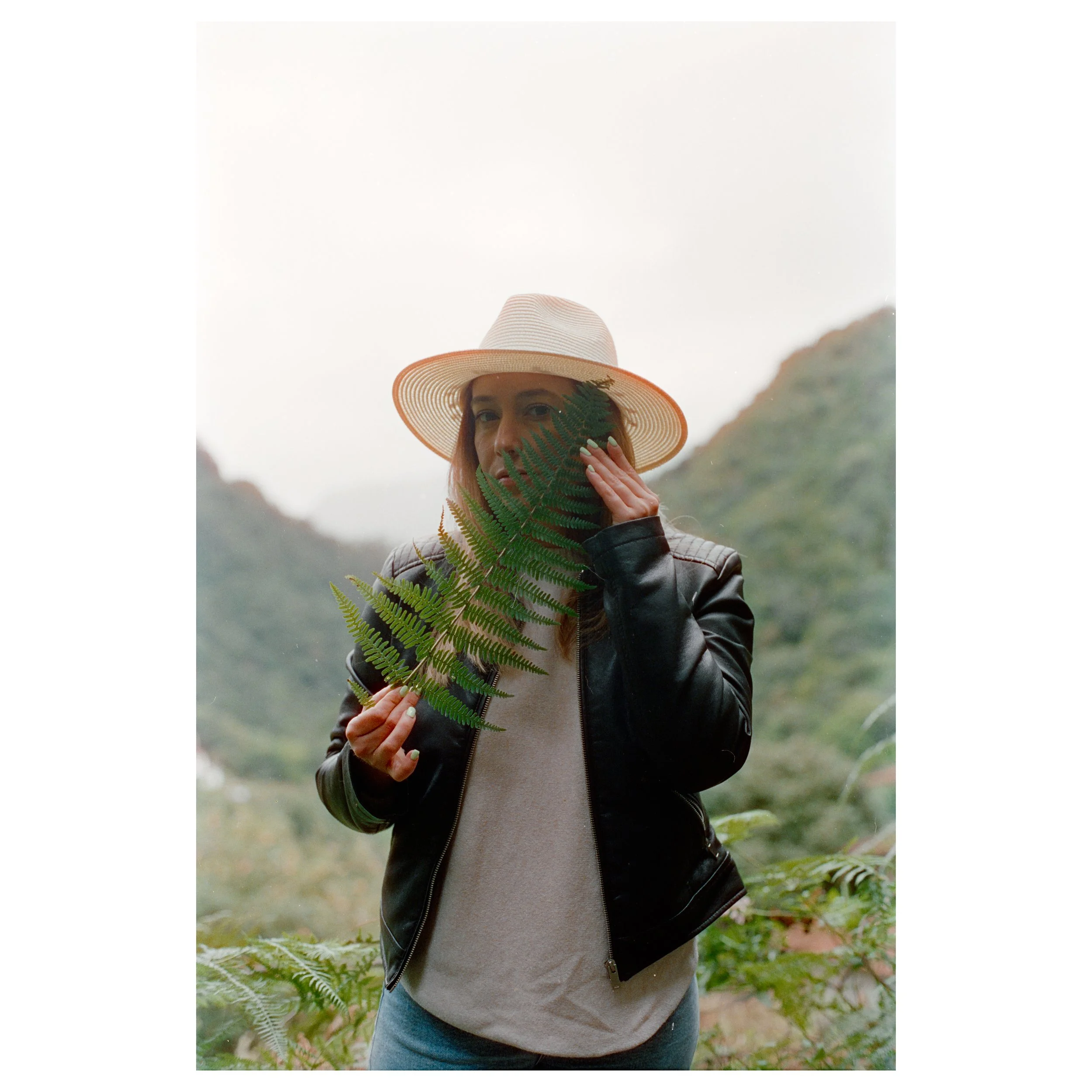 A woman wearing a wide-brimmed straw hat, a black leather jacket, and a beige top, holding a green fern leaf in front of her face with a misty mountainous landscape in the background.