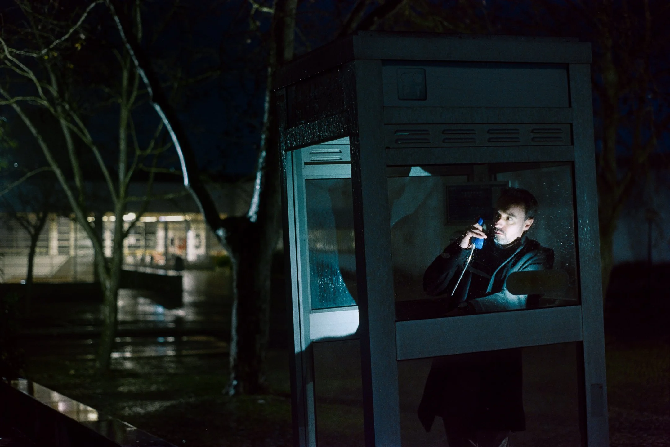 A man stands inside a glass phone booth at night, speaking on a phone, with rain on the glass and trees in the background.