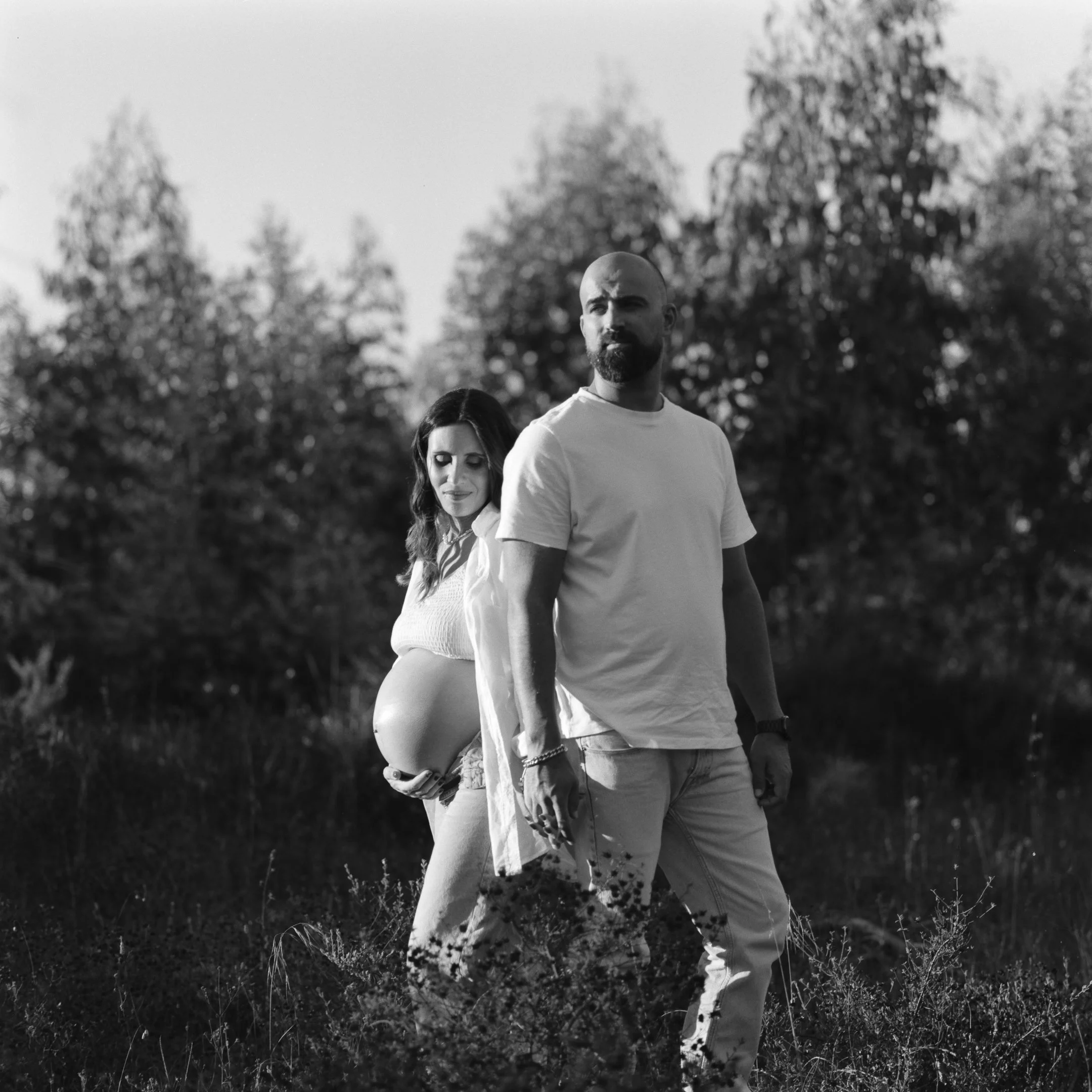 A pregnant woman standing outdoors in a field with a man, both looking towards the camera. The woman is behind the man, with her hand on her belly.