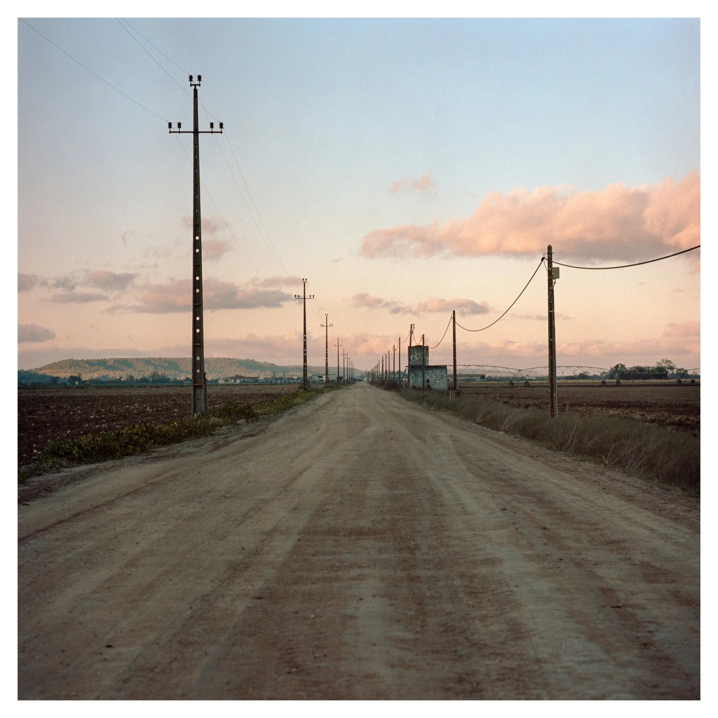 A rural dirt road with utility poles and wires on both sides, leading towards distant hills under a partly cloudy sky at sunset.