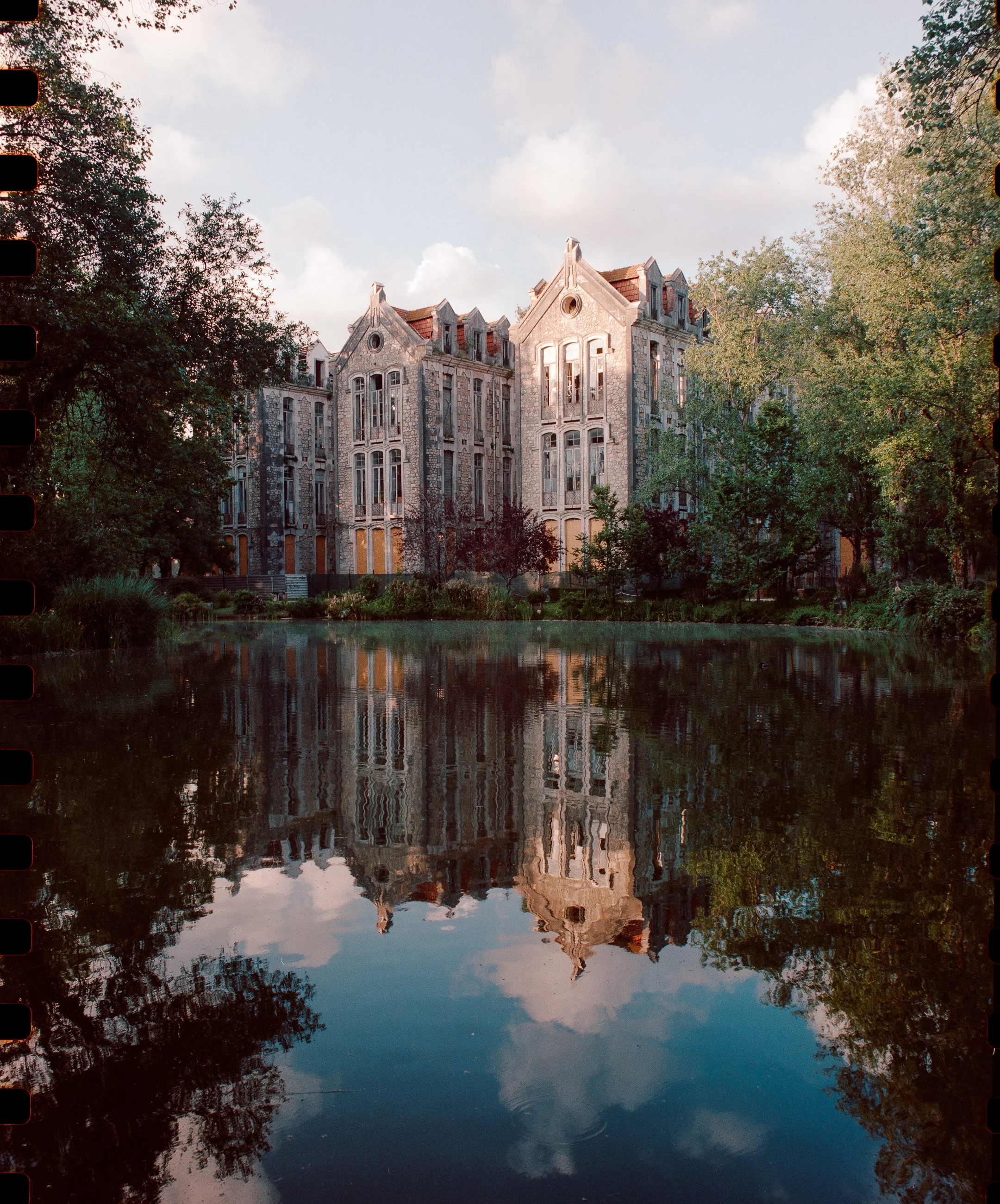 A large, old stone mansion with steep gabled roofs and numerous windows, reflected in a calm pond surrounded by trees. photo by Bruno Novais