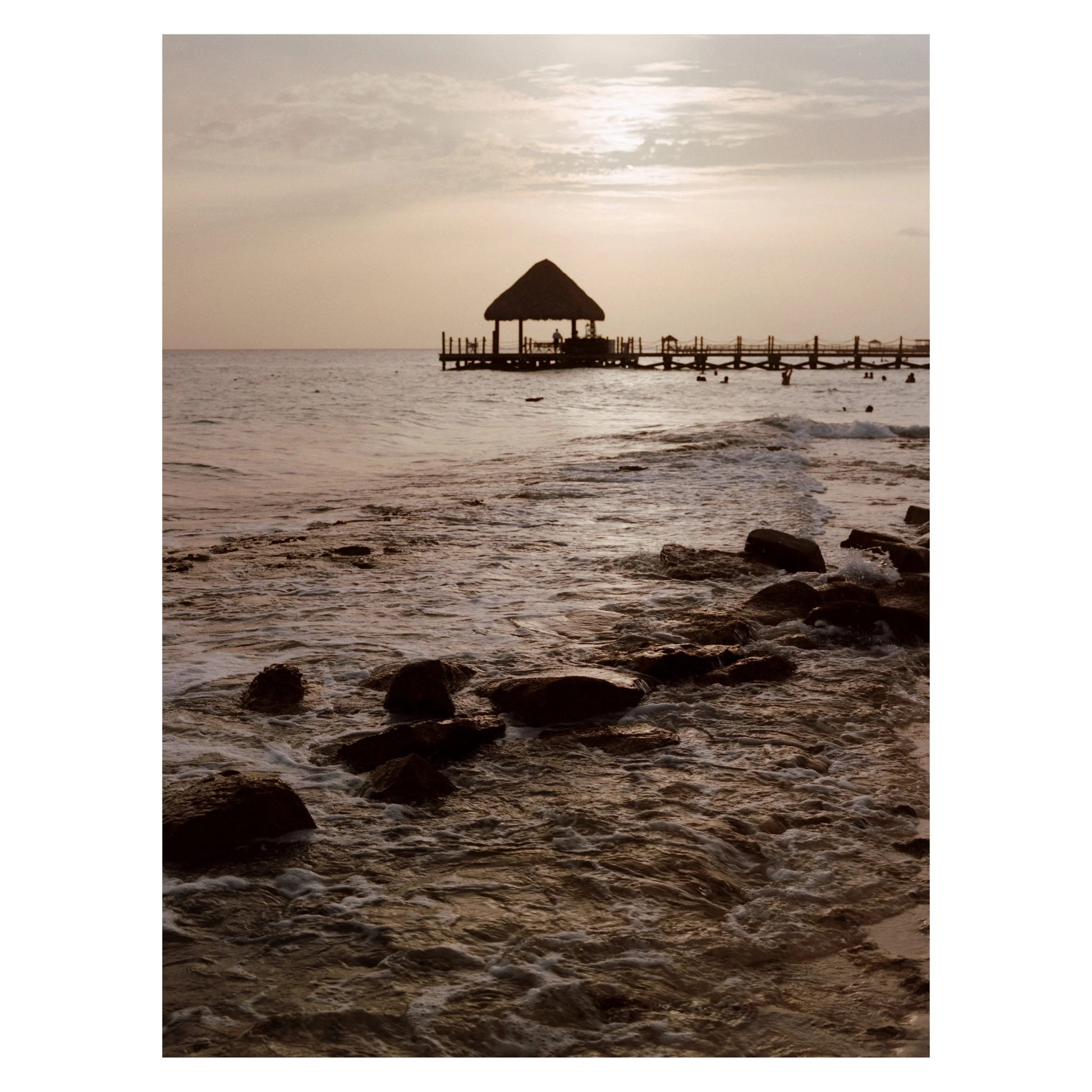 Sunset over the ocean with a thatched-roof pier in the background and rocks in the shallow water in the foreground.