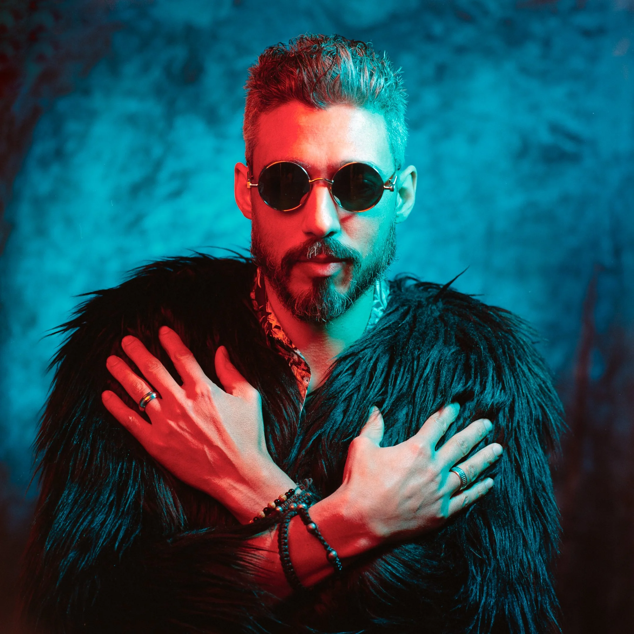 A man with short hair and a beard wearing round sunglasses and a black faux fur coat, hugging himself against a blue-toned background.